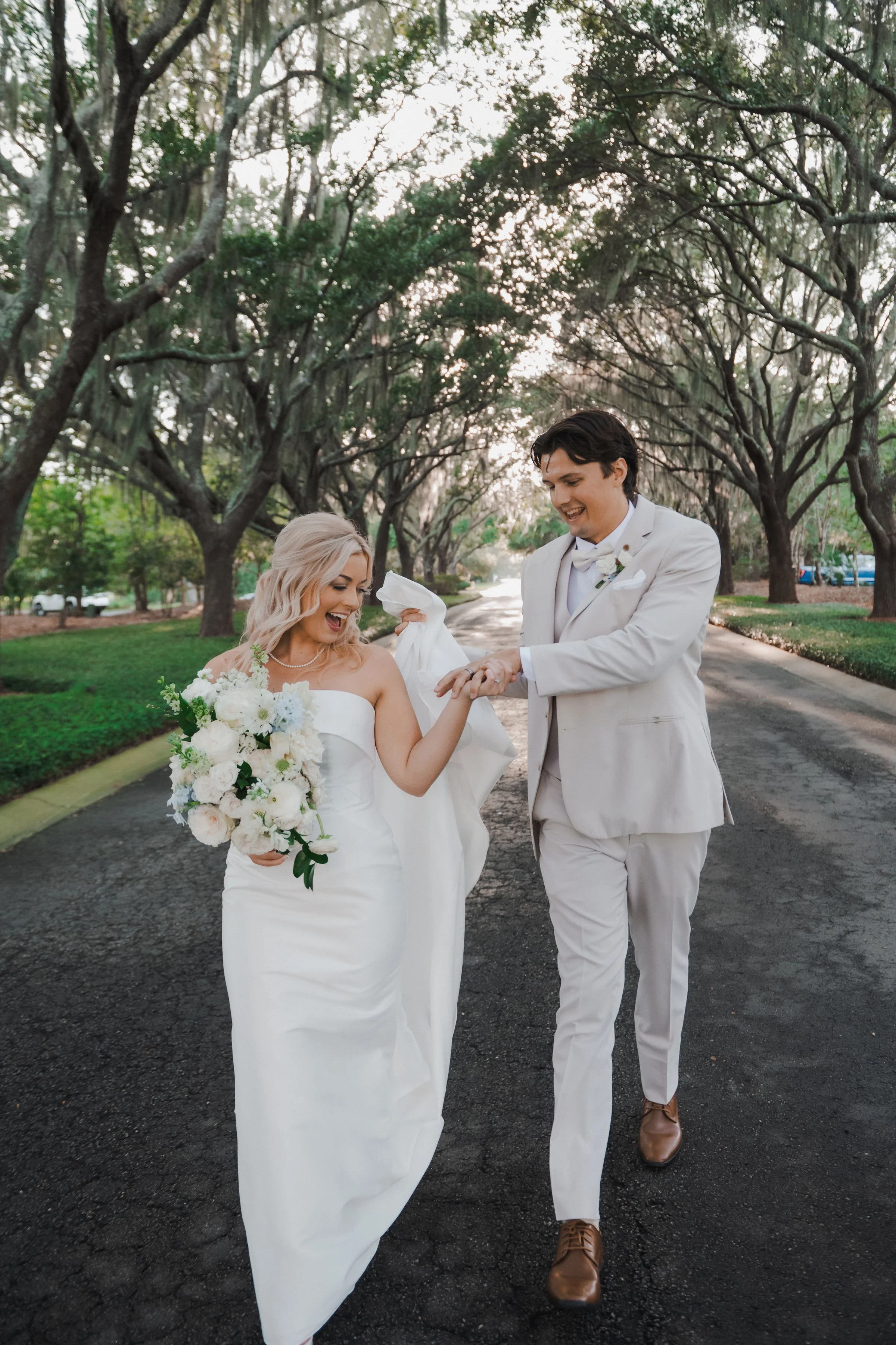 A bride and groom walking on a tree-lined street during their wedding day, with the bride holding a bouquet of white flowers and the groom looking at her ring.