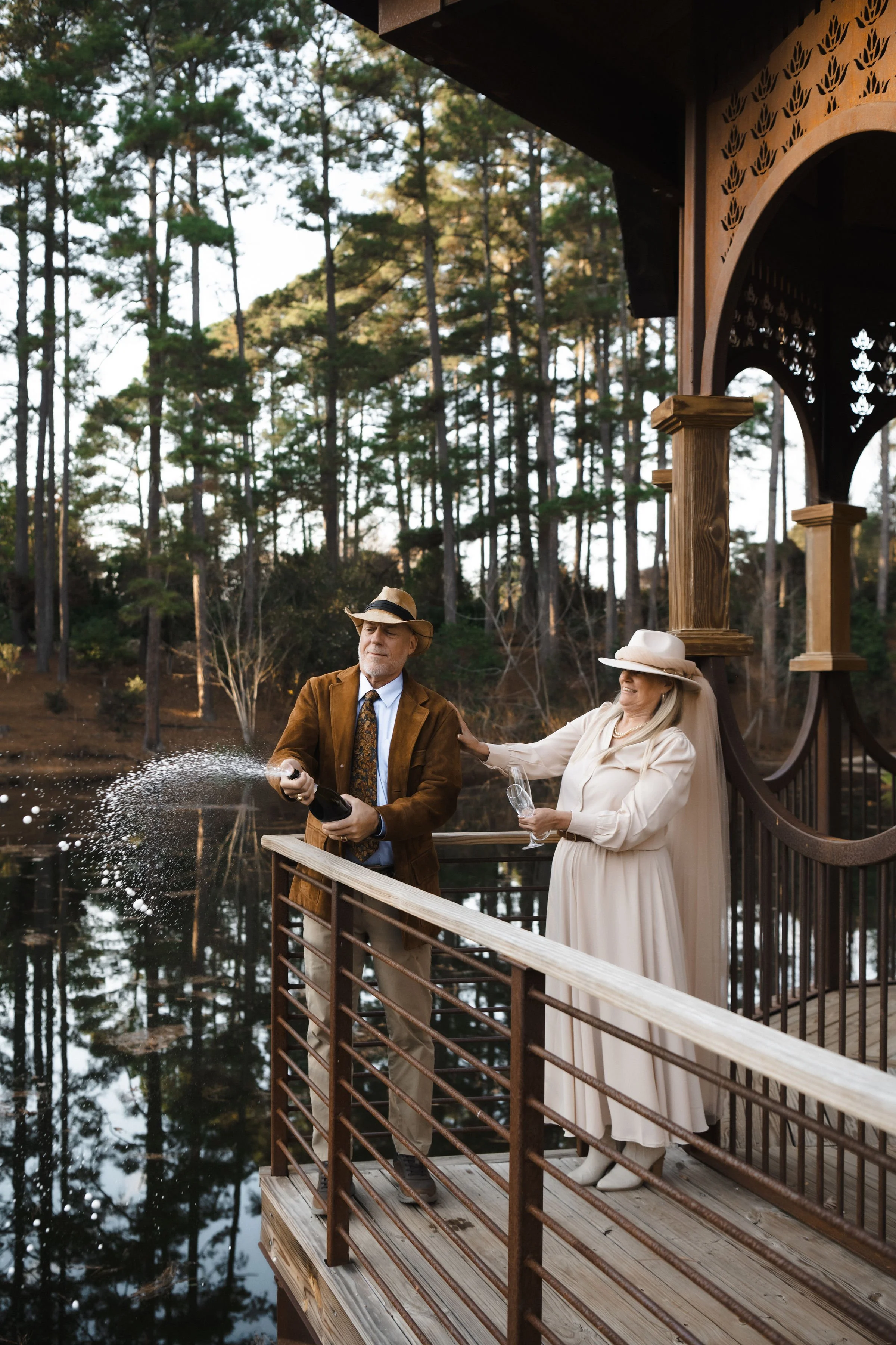 An elderly man in a brown jacket and a woman in a beige dress and hat celebrate on a wooden deck by a lake, with the man opening a bottle of champagne and the woman holding a glass, amidst a background of tall trees.