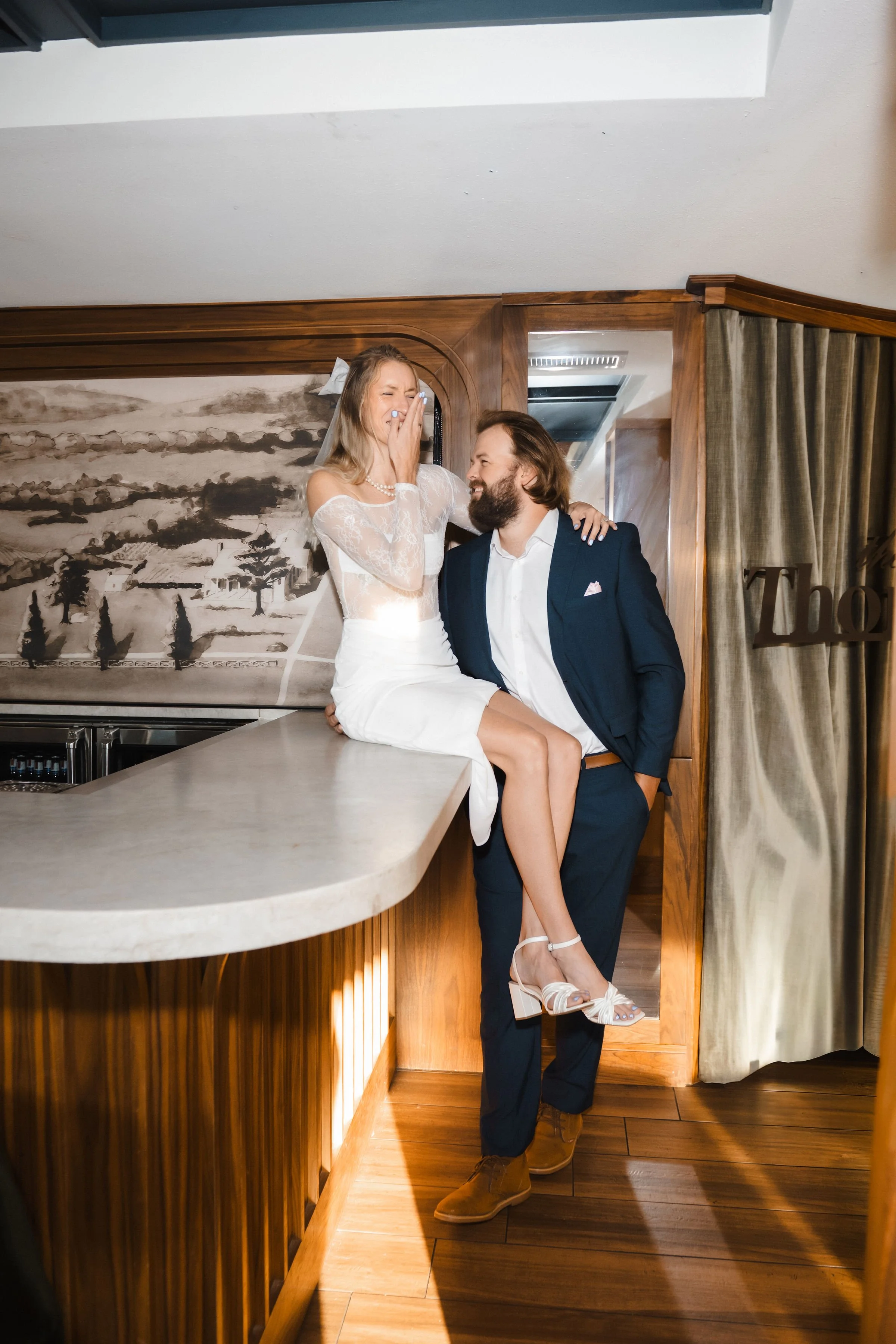 A couple celebrating a special moment indoors, with the woman sitting on a bar counter and the man standing beside her, both smiling and sharing a joyful moment.