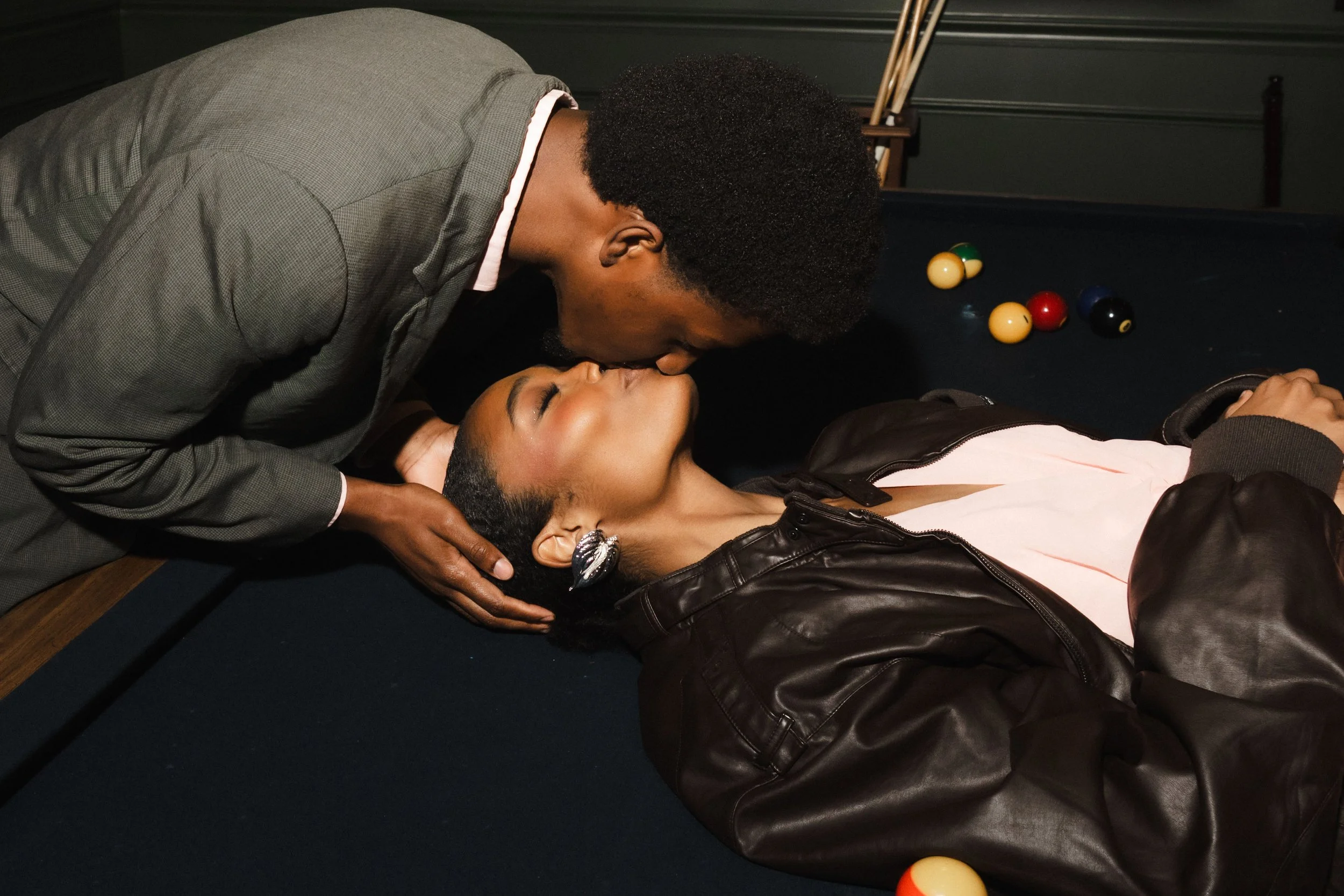 A man and woman share a kiss above a pool table, with billiard balls scattered nearby.