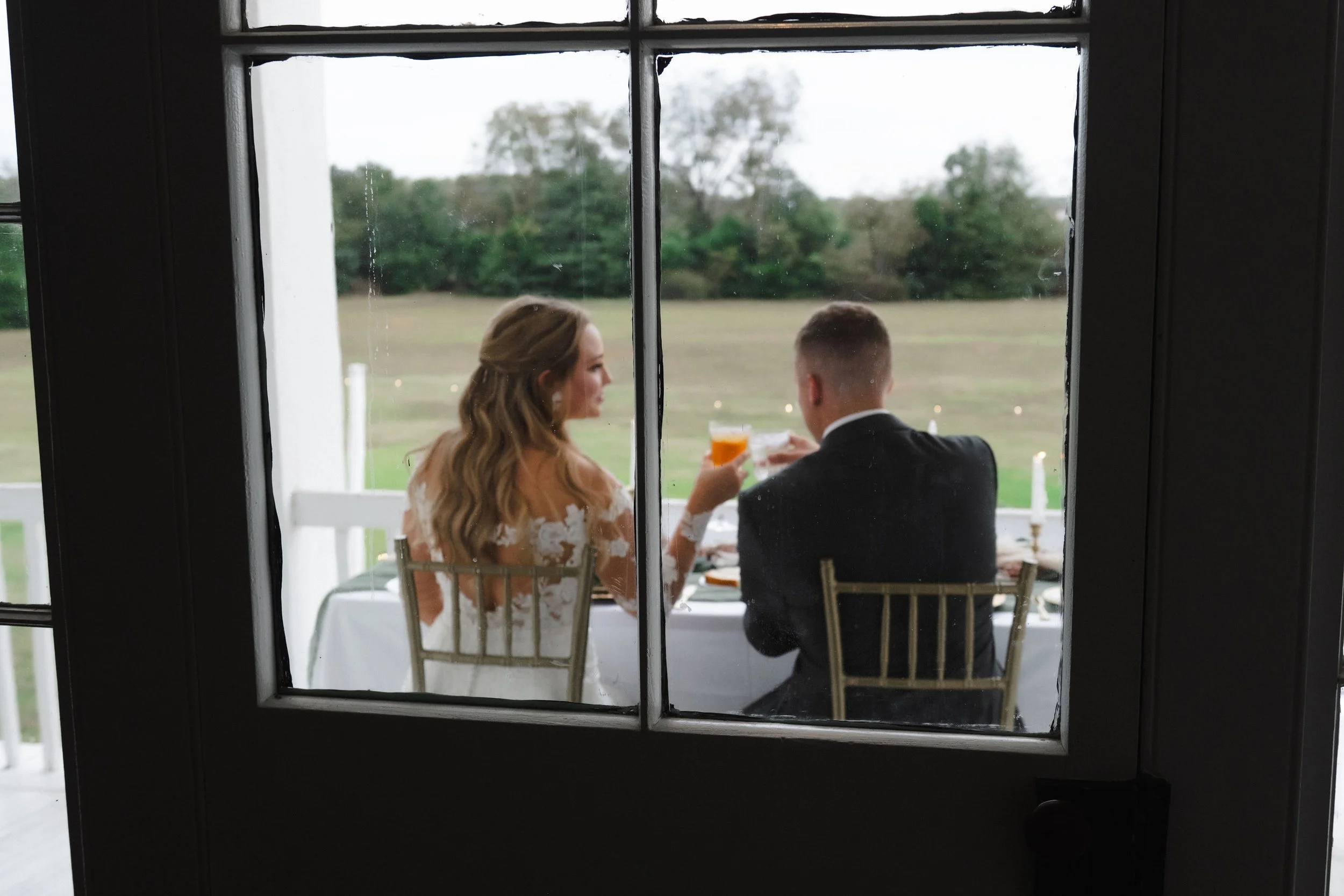 A bride and groom sitting at a table, holding glasses and sharing a toast, seen through a window with black framing, outdoors with green trees and open field in the background.