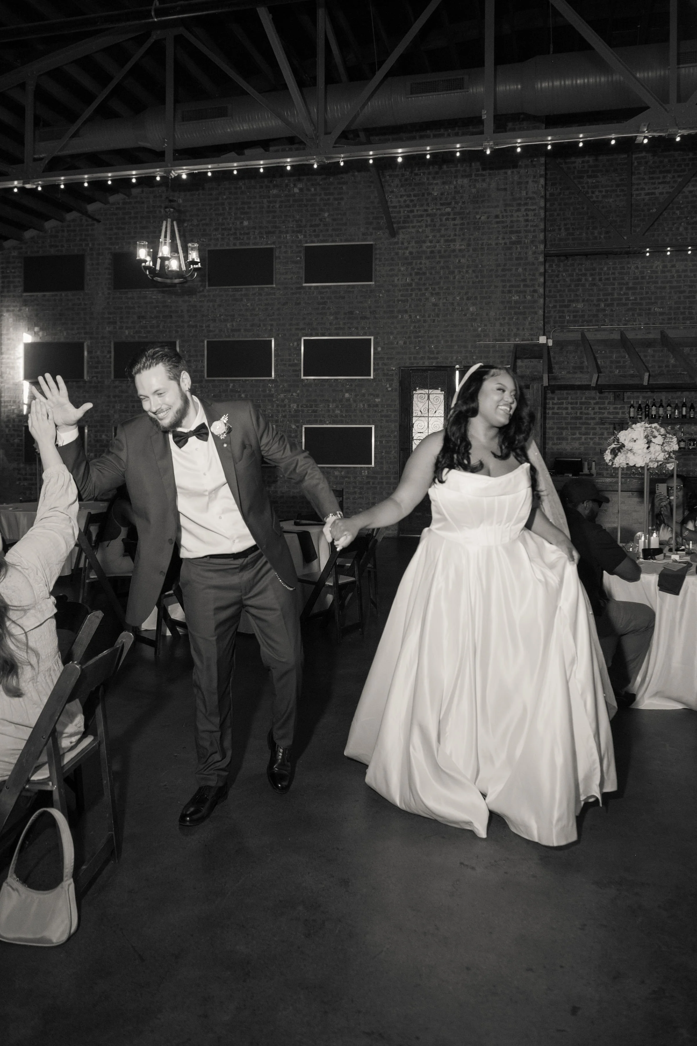 Black and white photo of a bride and groom dancing at their wedding reception, holding hands and smiling.