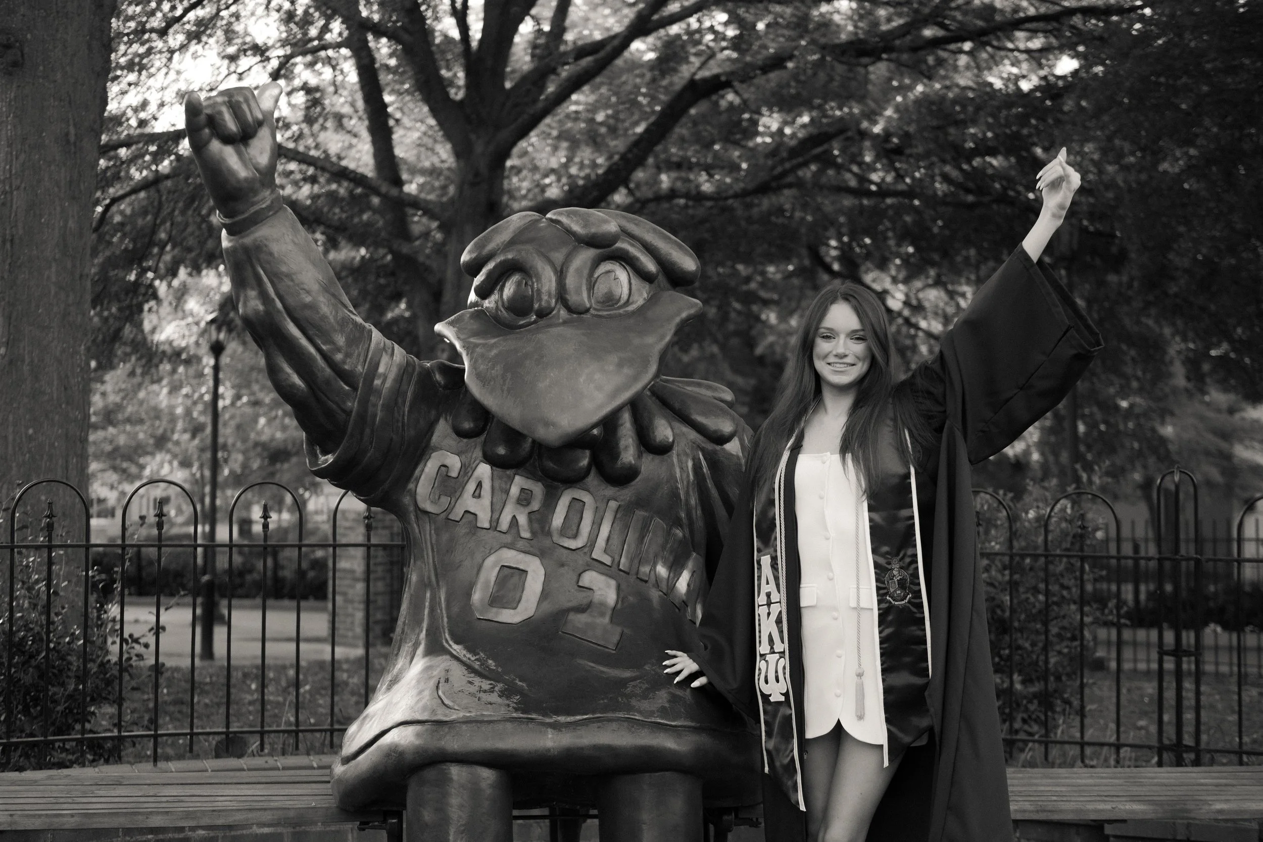 A young woman in a graduation gown standing next to a statue of a bird mascot with the word 'CAROLINA' and '01' on it, celebrating her graduation outdoors with trees and a fence in the background.