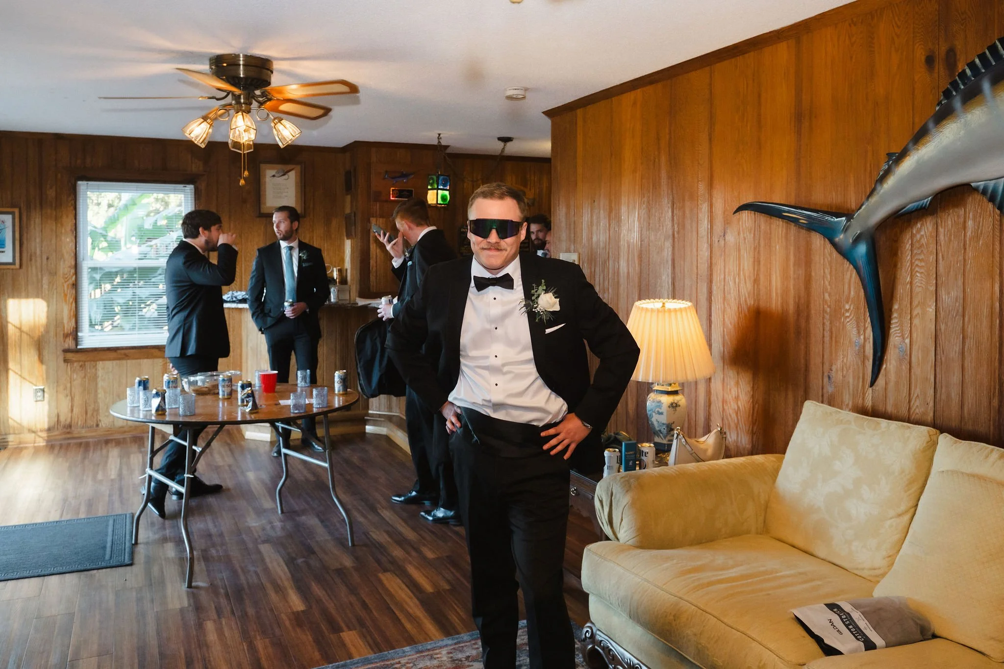 A man in a tuxedo with sunglasses, standing in a wood-paneled room with a yellow couch and a group of men in suits in the background, some talking and drinking, with a table of cans and glasses.