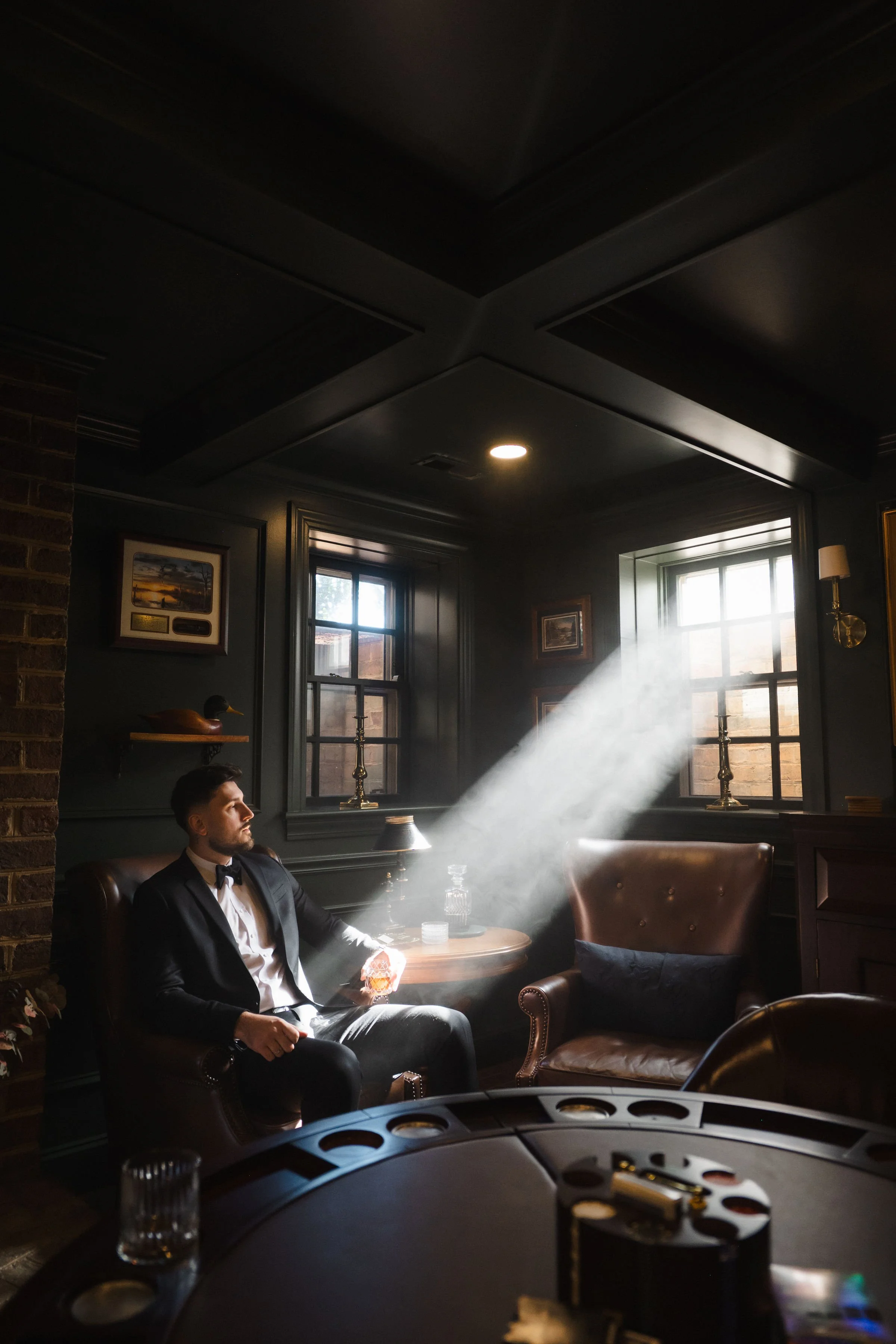 A man in a tuxedo sitting on a leather armchair in a dimly lit room with two windows, sunlight streaming through, casting a beam of light across the room. 