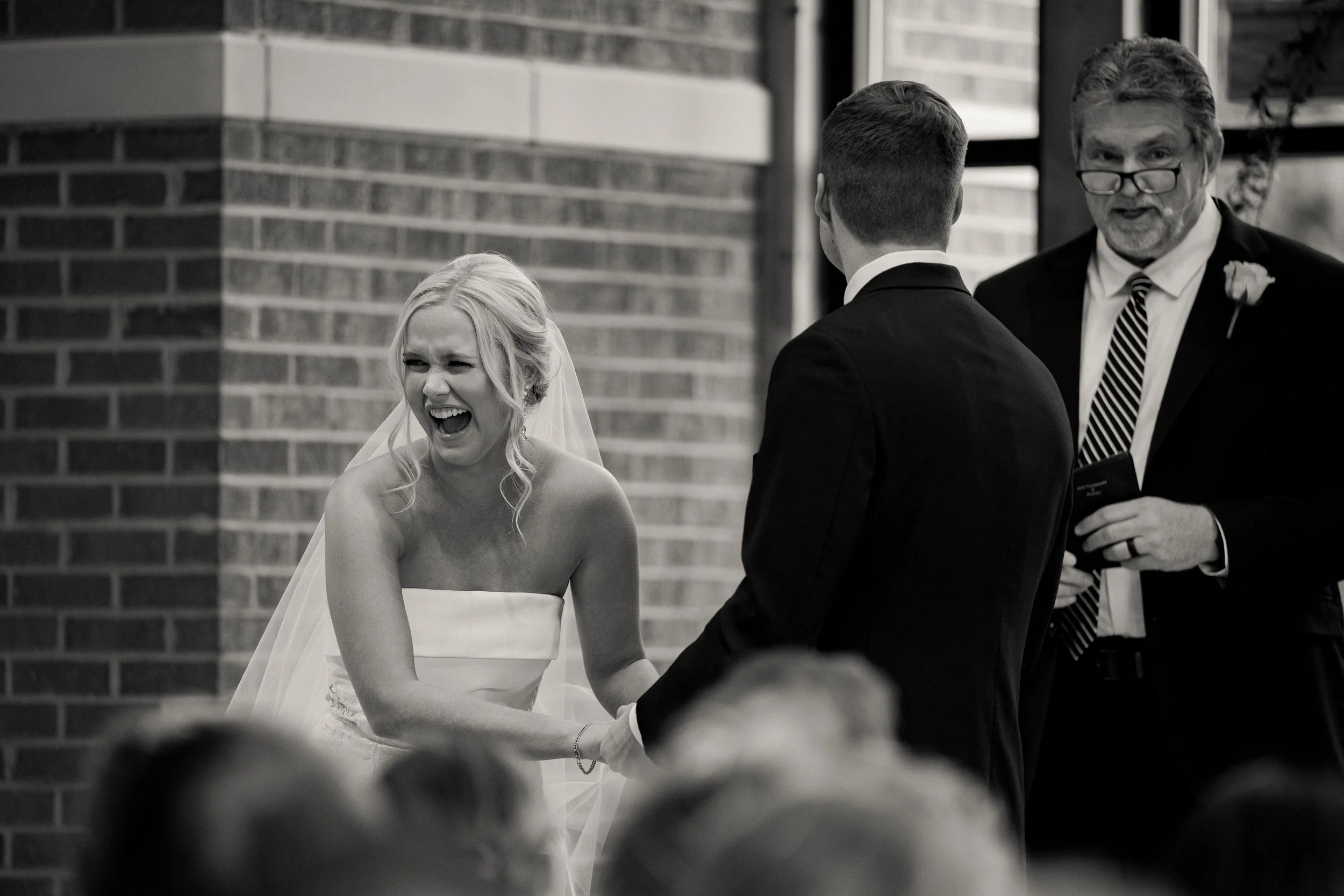 A bride and groom exchanging vows at their wedding ceremony inside a church, with an officiant standing nearby. The bride is laughing joyfully while holding the groom's hand.