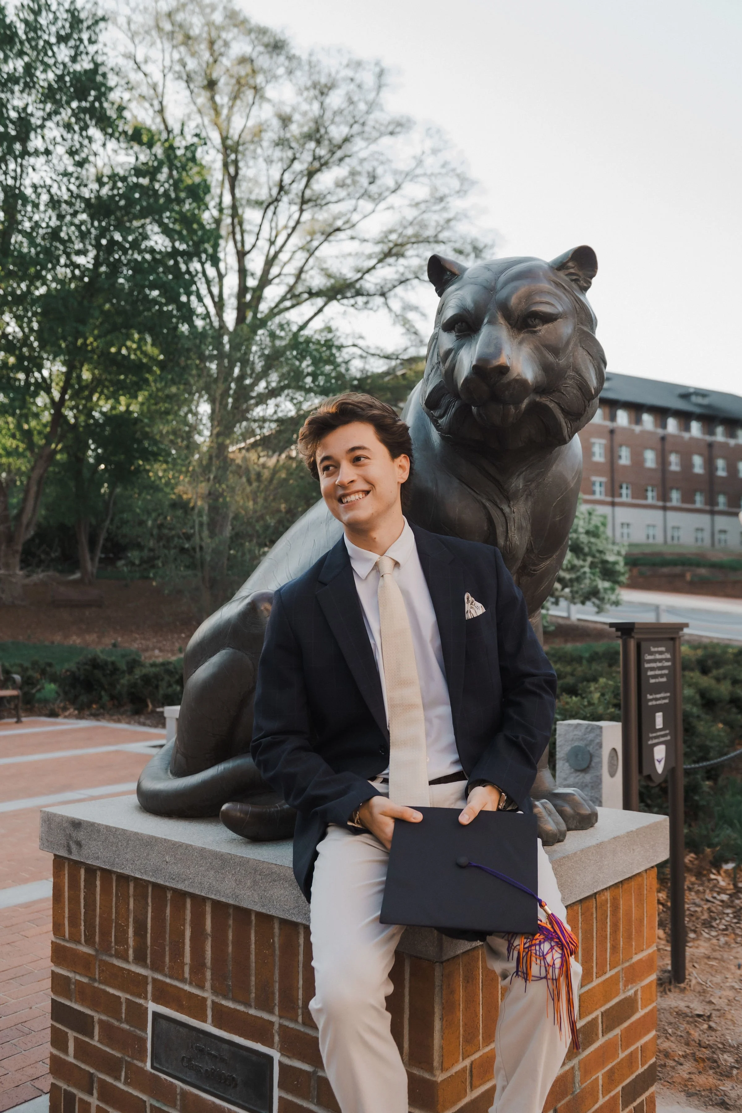 A young man in a suit sitting on a brick pedestal in front of a large lion statue, outdoors during late afternoon, holding a graduation cap with a colorful tassel, smiling.