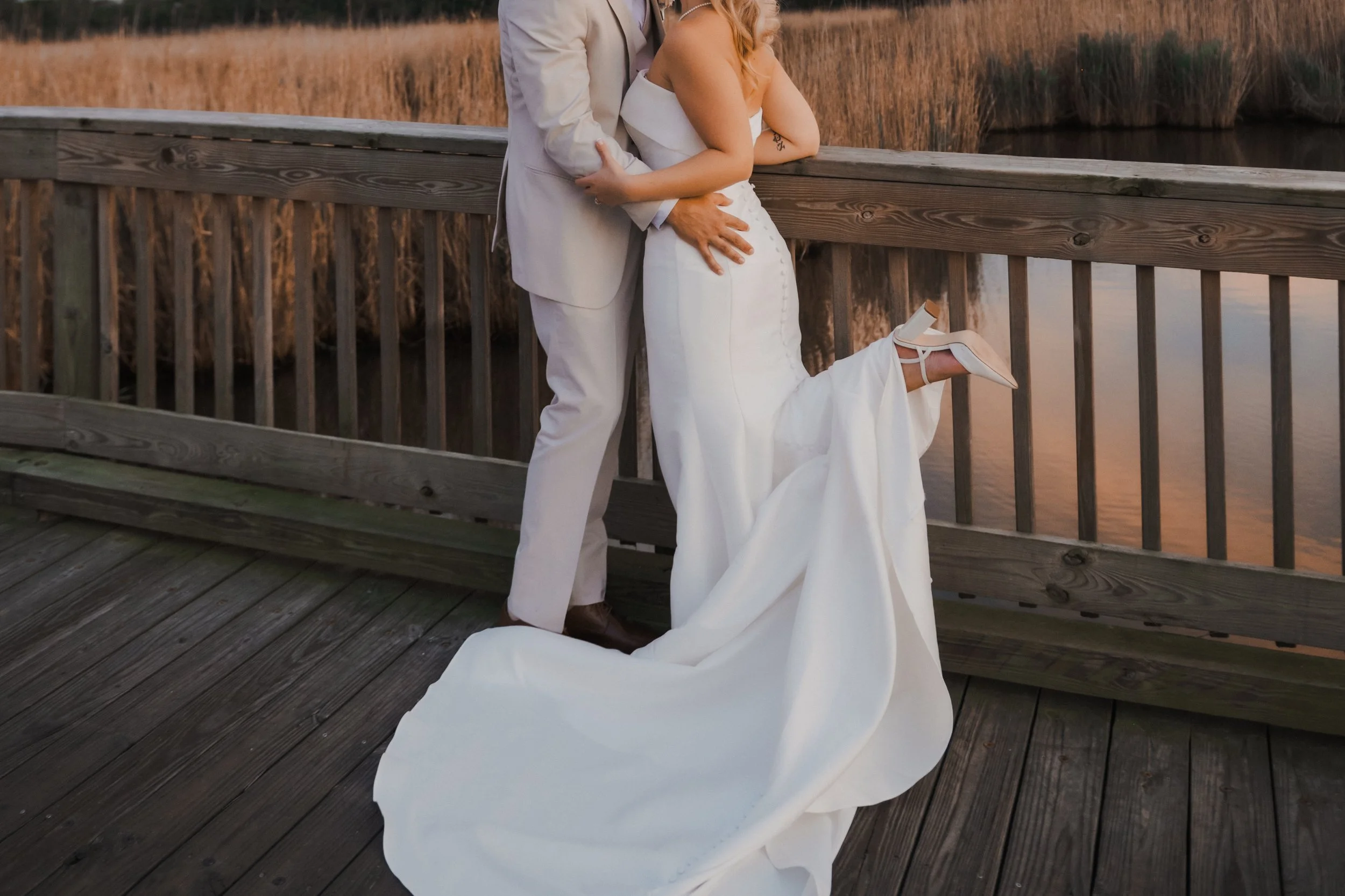 A couple dressed in white wedding attire standing on a wooden bridge by a river at sunset, with the woman lifting one foot to show her white high heel.