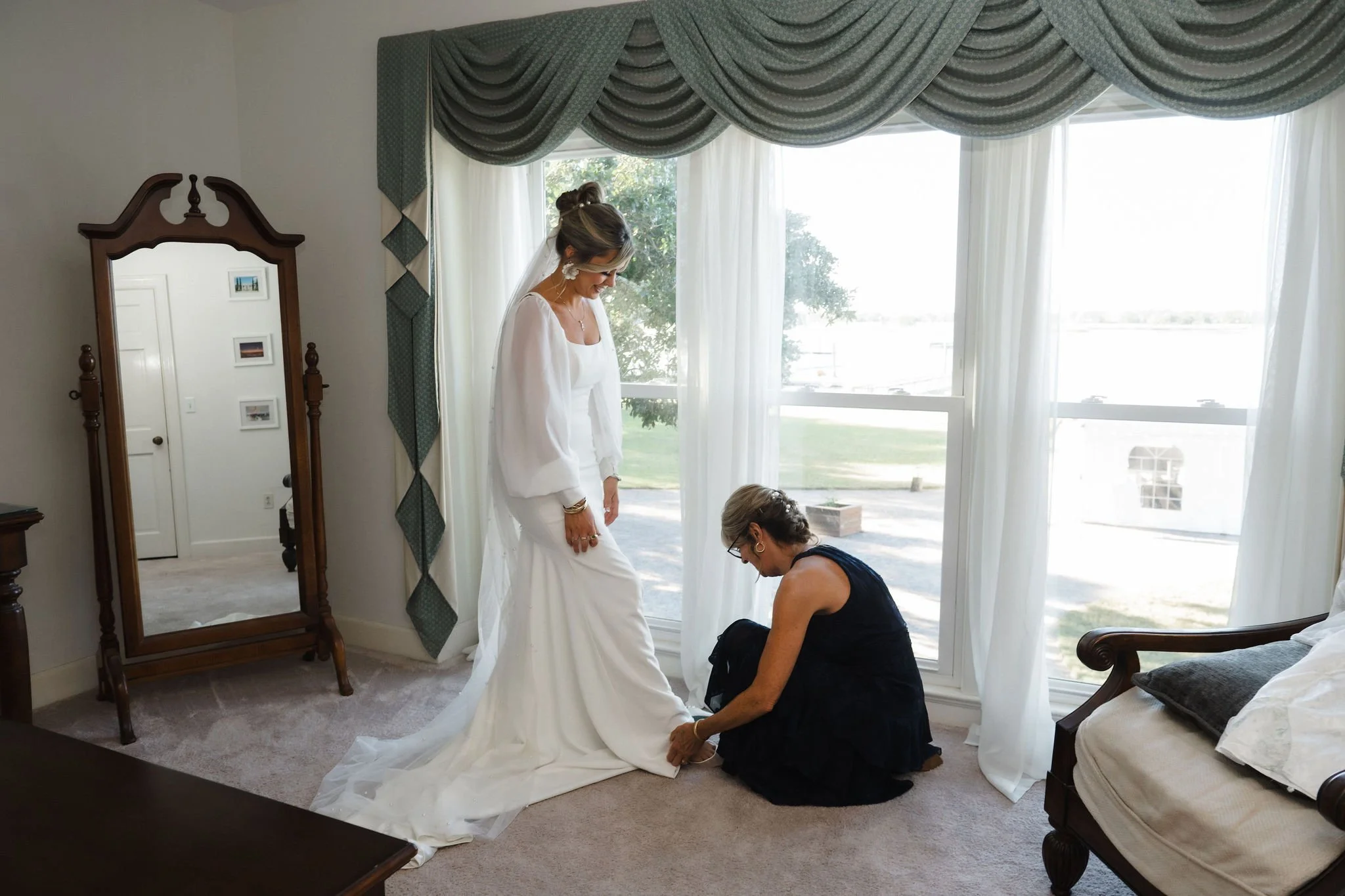 A bride in a white wedding dress is getting her shoes tied by a woman kneeling on the carpeted floor near a large window with green and white curtains.