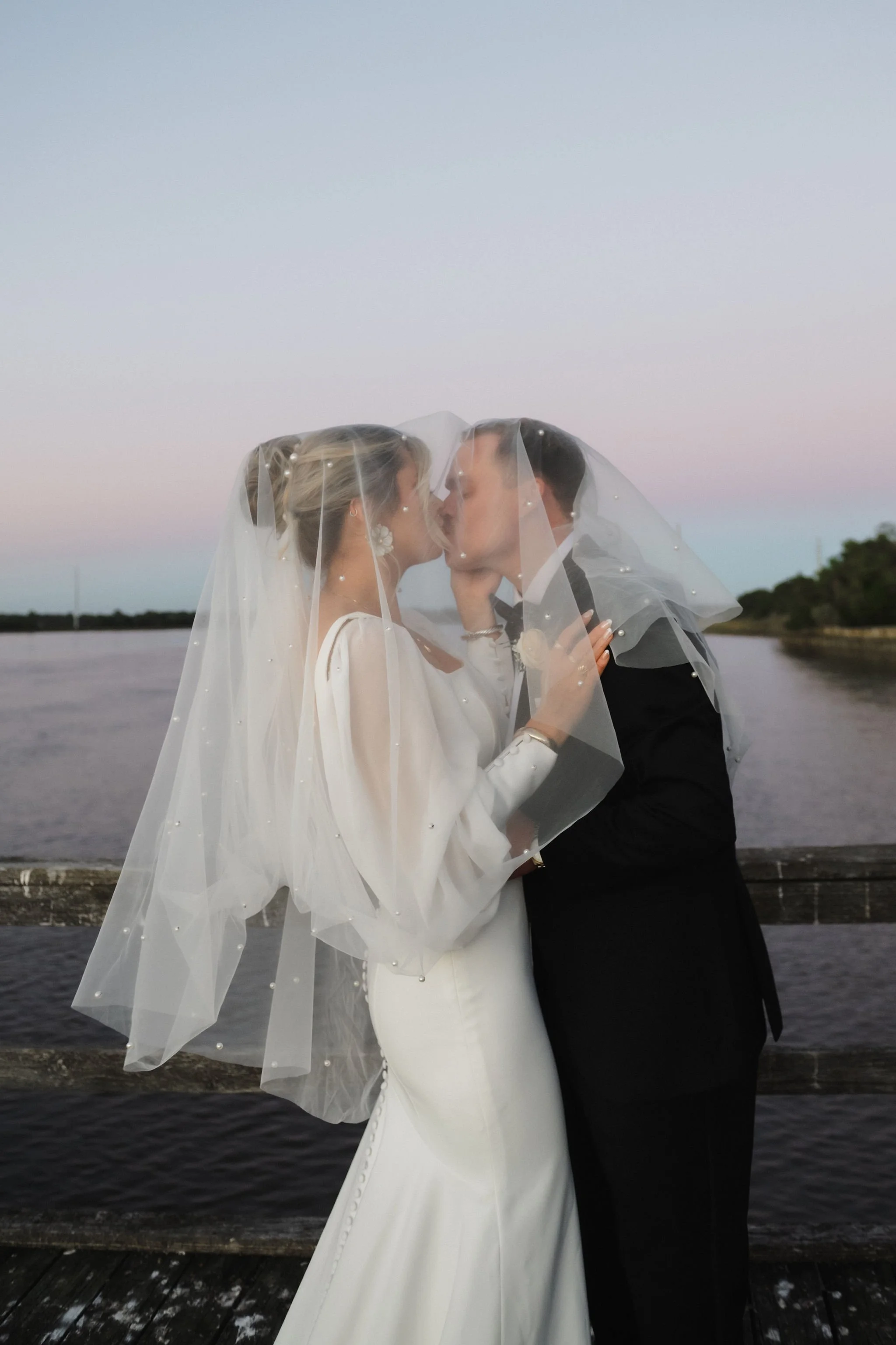 A bride and groom kissing under a veil on a dock by the water at sunset.