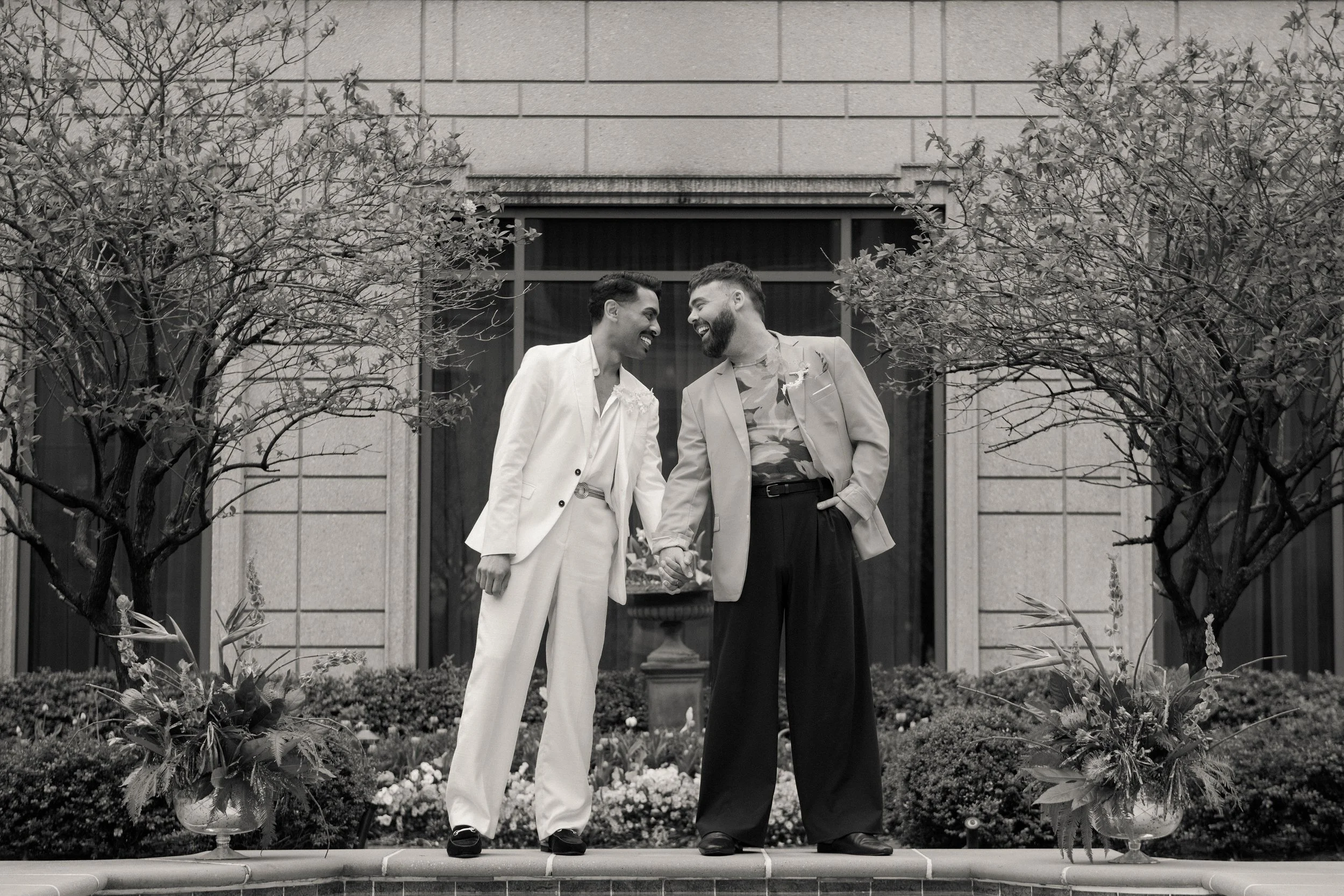 Two men holding hands and smiling at each other in front of a building, with trees and flower arrangements on either side of a fountain