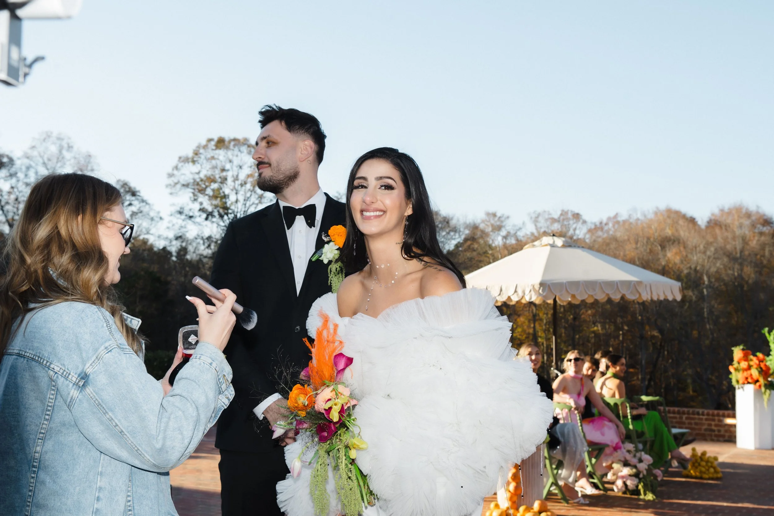 A bride and groom at their outdoor wedding ceremony, with the bride in a white dress holding a flower bouquet, and the groom in a tuxedo with a boutonniere, smiling as a woman in glasses and a denim jacket uses a microphone near them. Guests are seat