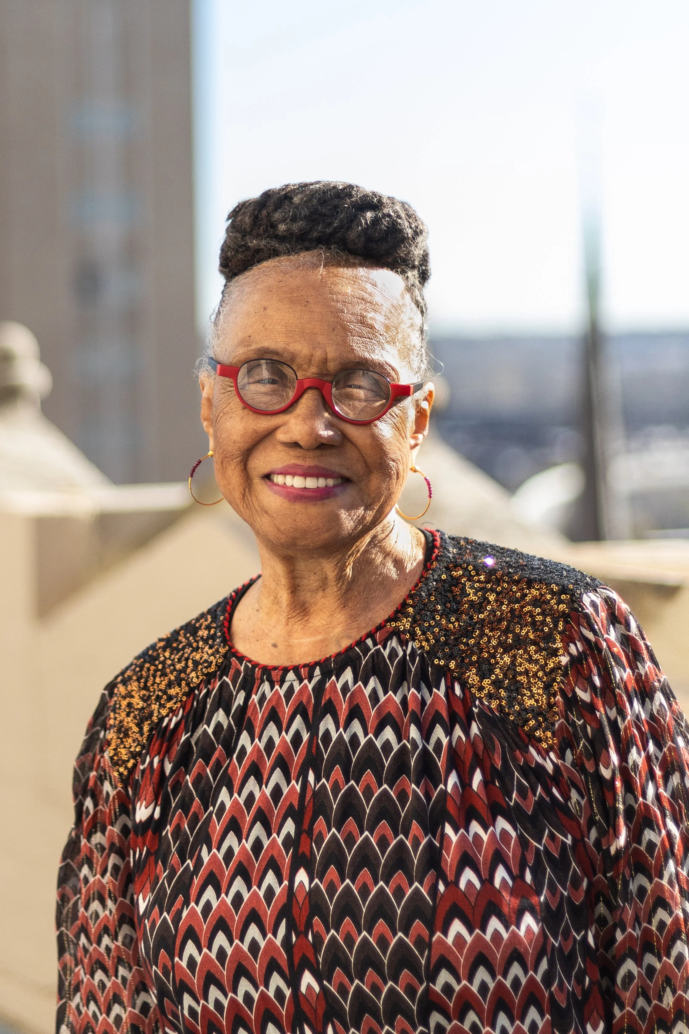 Portrait of an older woman with short gray hair, wearing glasses, a teal blazer, and a blue floral blouse, against a plain white background.