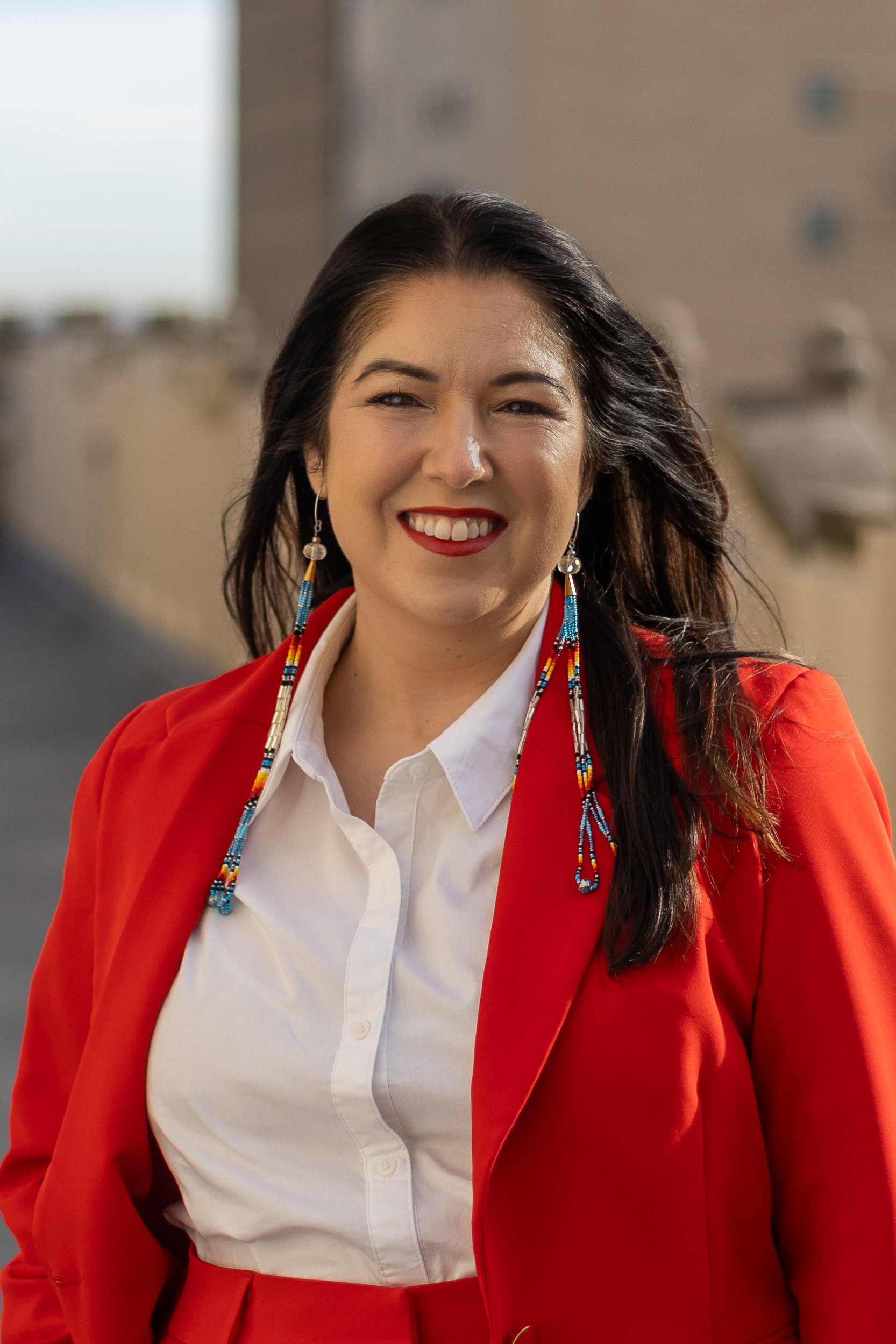 A woman with shoulder-length brown hair wearing a black blazer, black top, and gold belt, accessorized with colorful earrings, smiling against a plain white background.