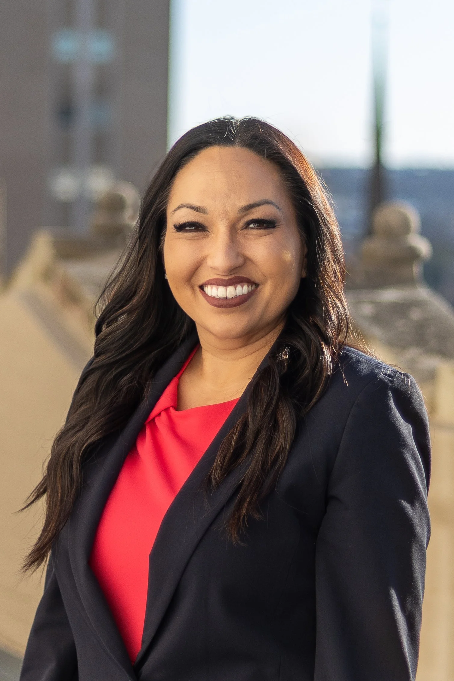 Portrait of a woman with dark, wavy hair, smiling, wearing a red blazer and black top, standing against a plain white background.