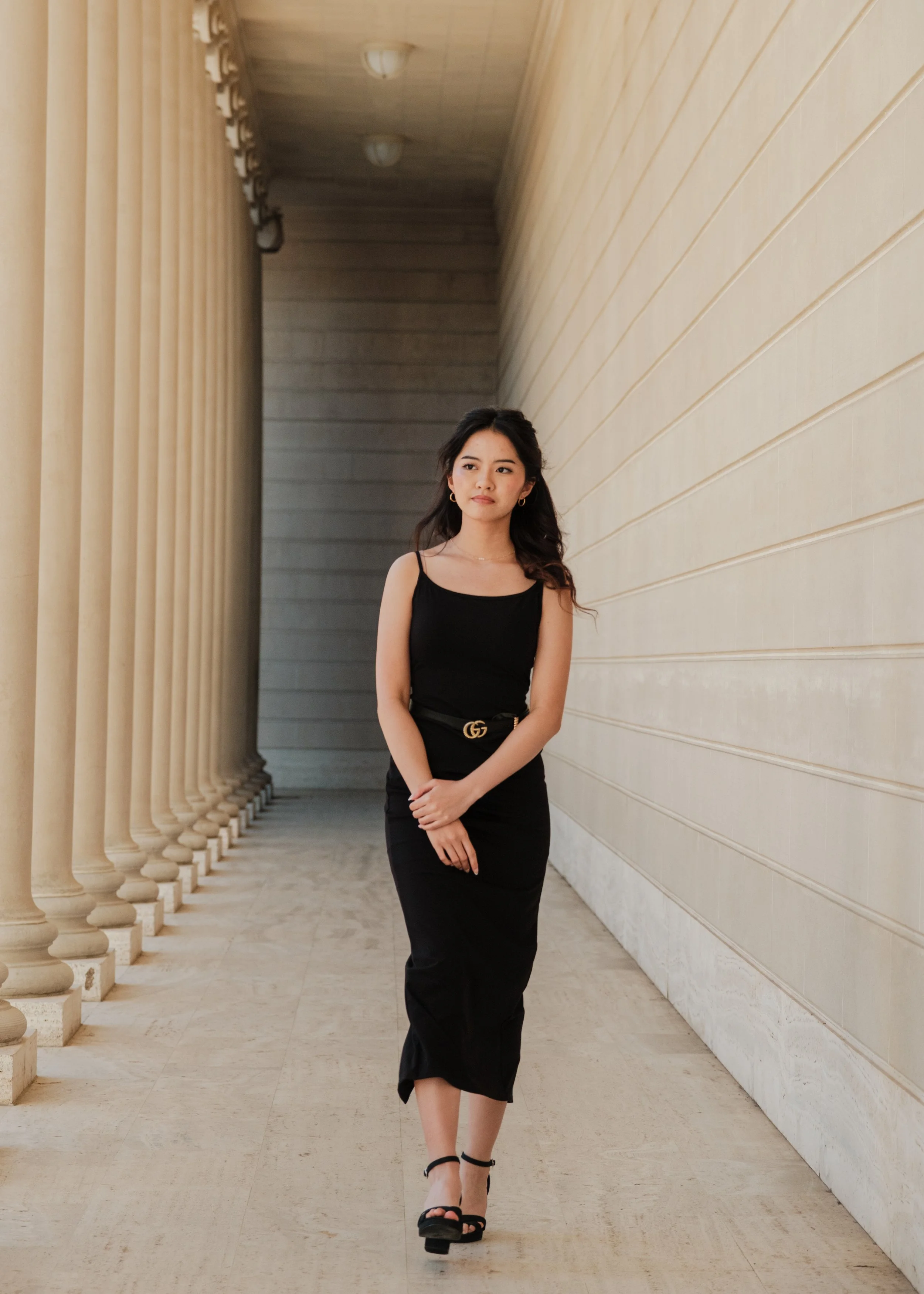 A woman wearing a black dress with spaghetti straps and black high-heeled sandals walking along a corridor with tall columns on one side and a textured beige wall on the other.