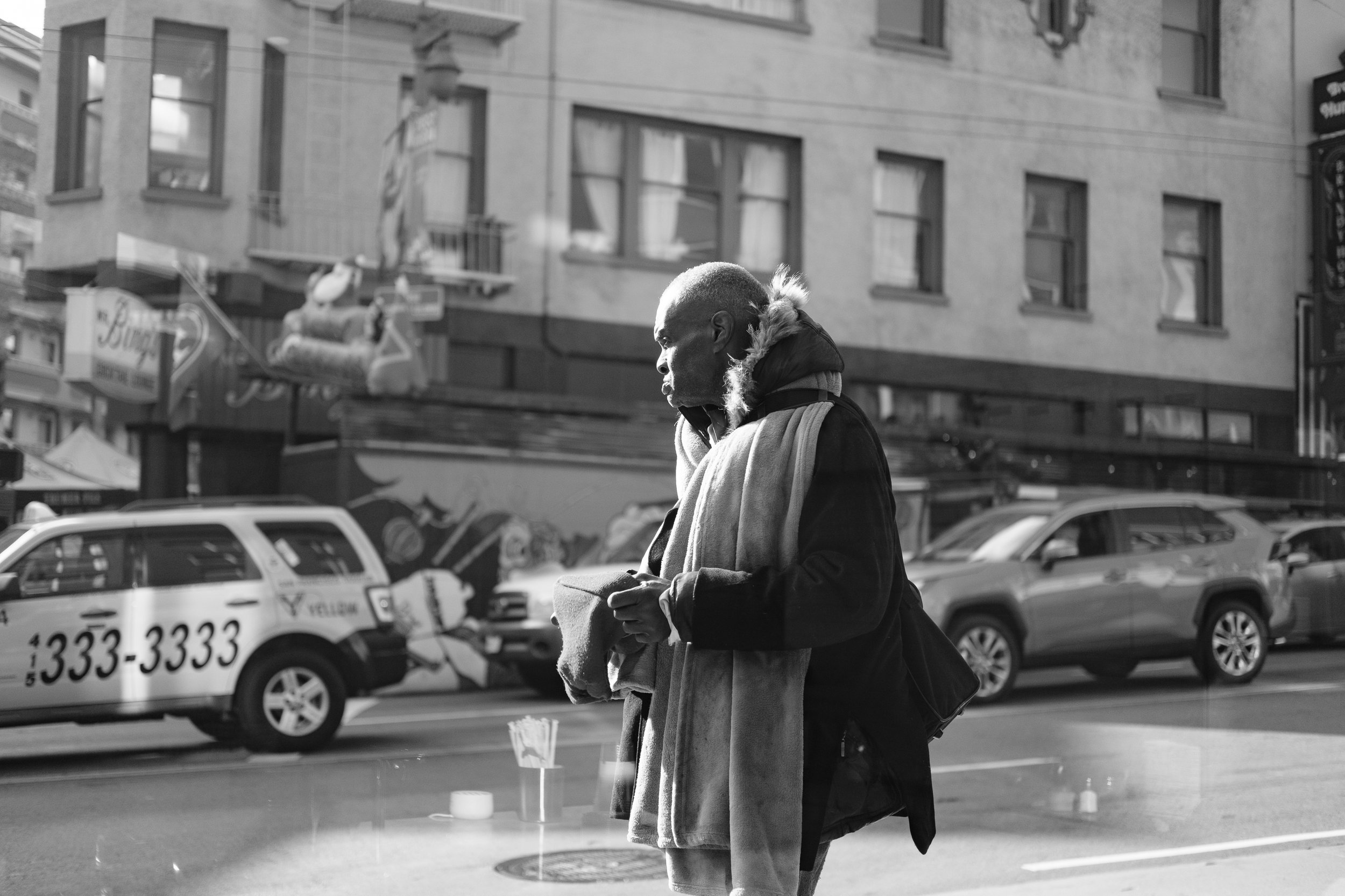 A black and white photo of an older man standing on a city sidewalk. He is dressed warmly with a scarf and jacket, and he appears to be looking at his phone. There are cars and a building with multiple windows in the background.