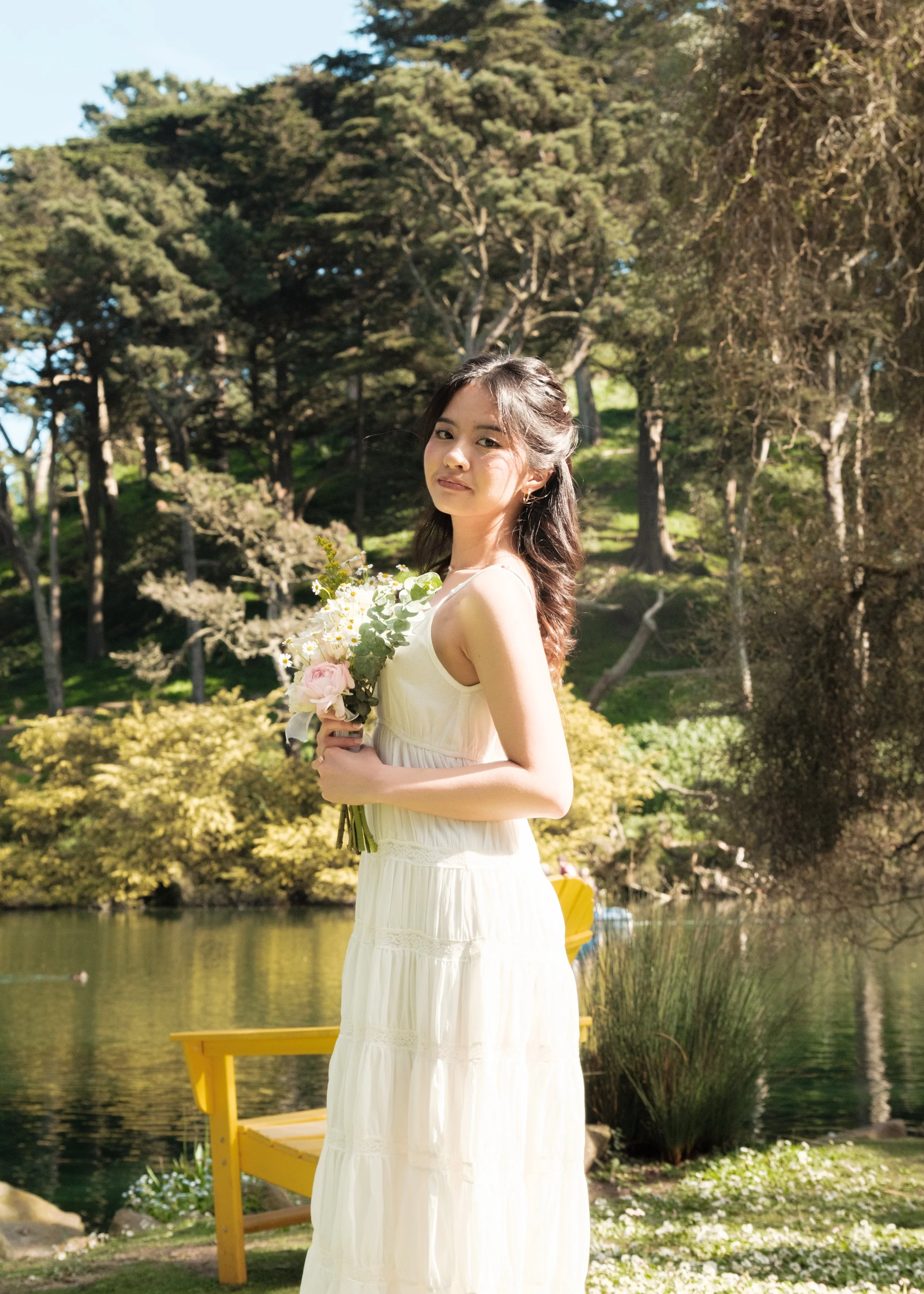 A woman in a white dress holding a bouquet of flowers, standing outdoors near a lake with trees in the background.