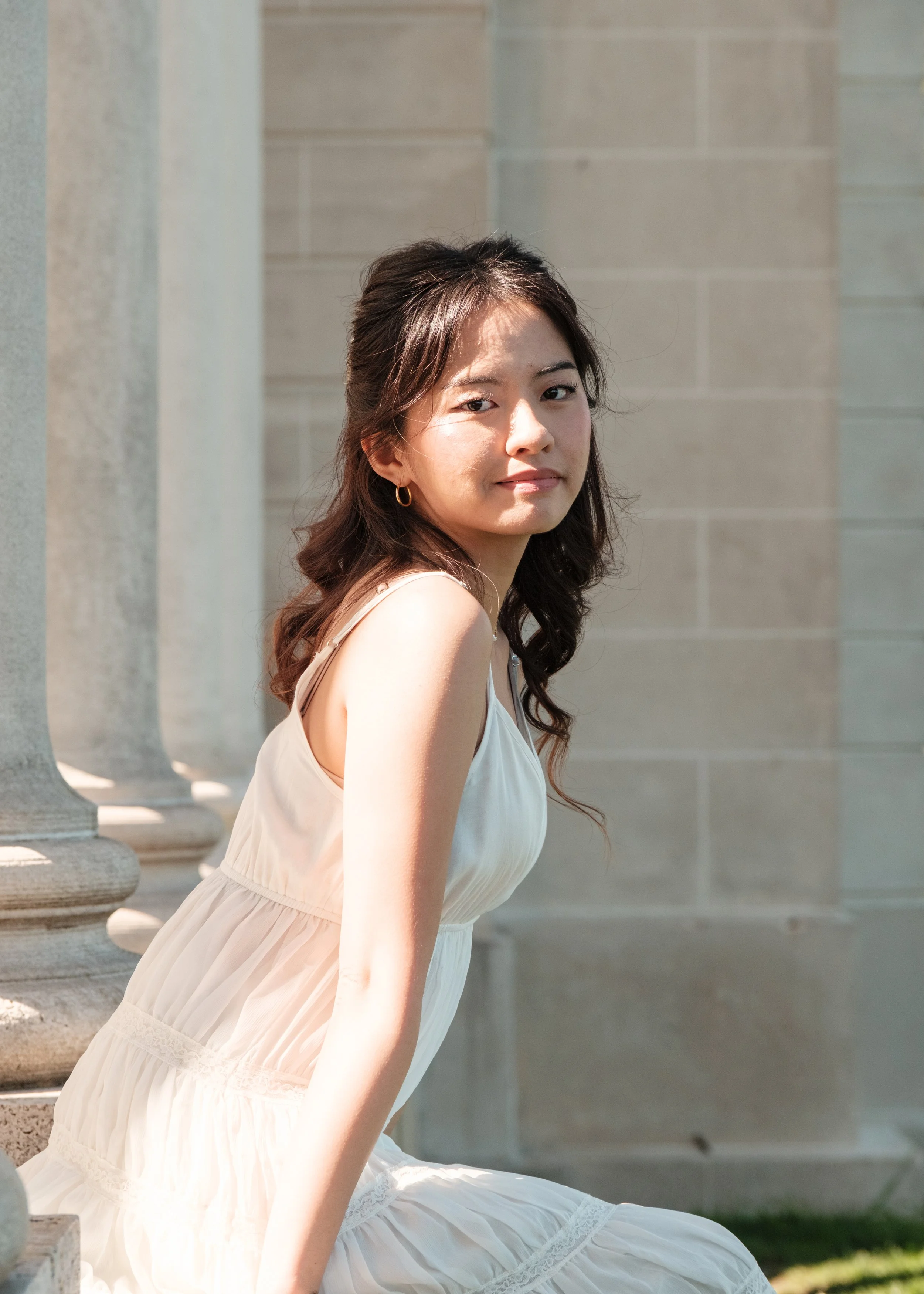 A young woman with long wavy hair and earrings, wearing a light-colored dress, sitting outdoors near stone columns and a stone wall, with sunlight illuminating her face.
