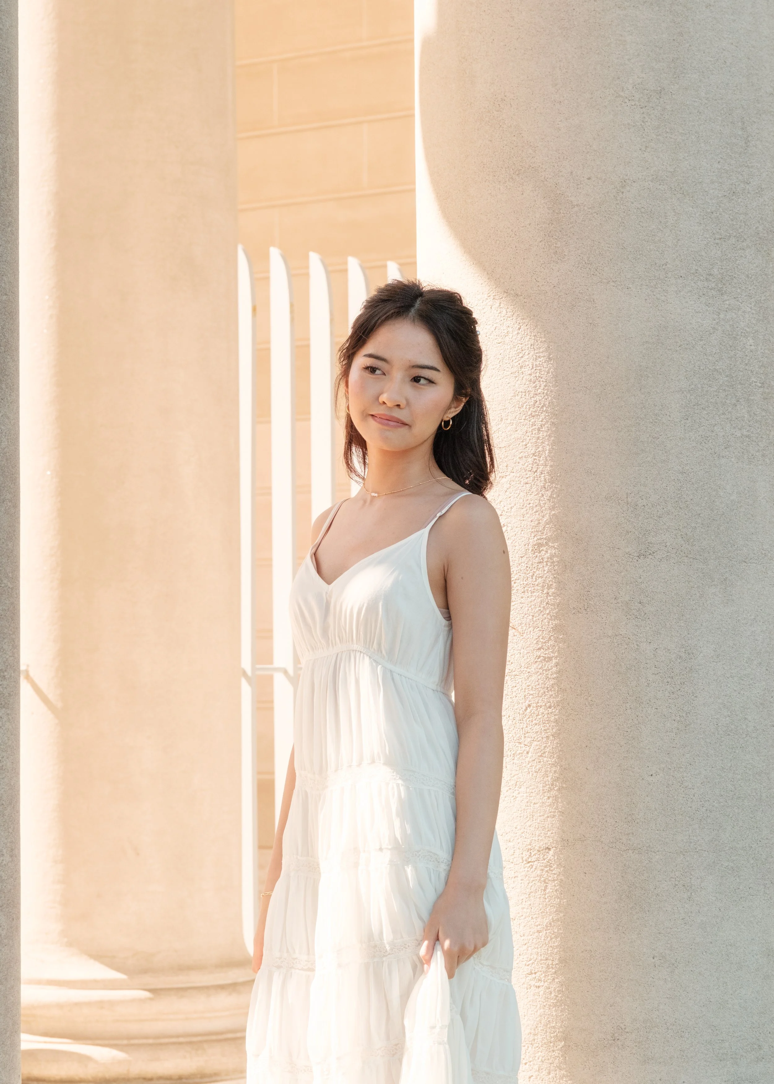 A young woman in a flowing white dress standing outdoors near large columns, looking thoughtfully to the side with sunlight on her face.