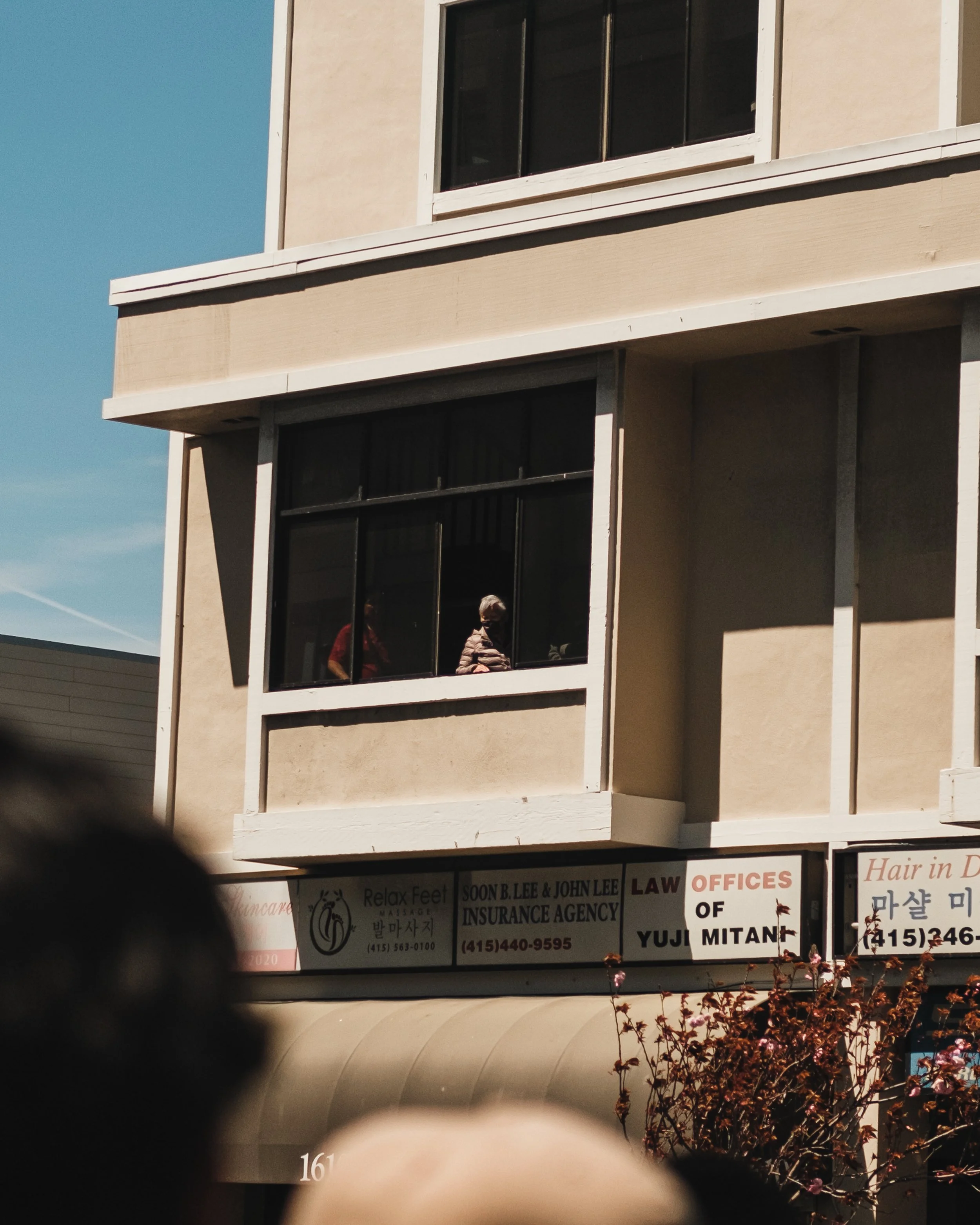 People looking out of a large apartment window in a multi-story building with storefronts below.
