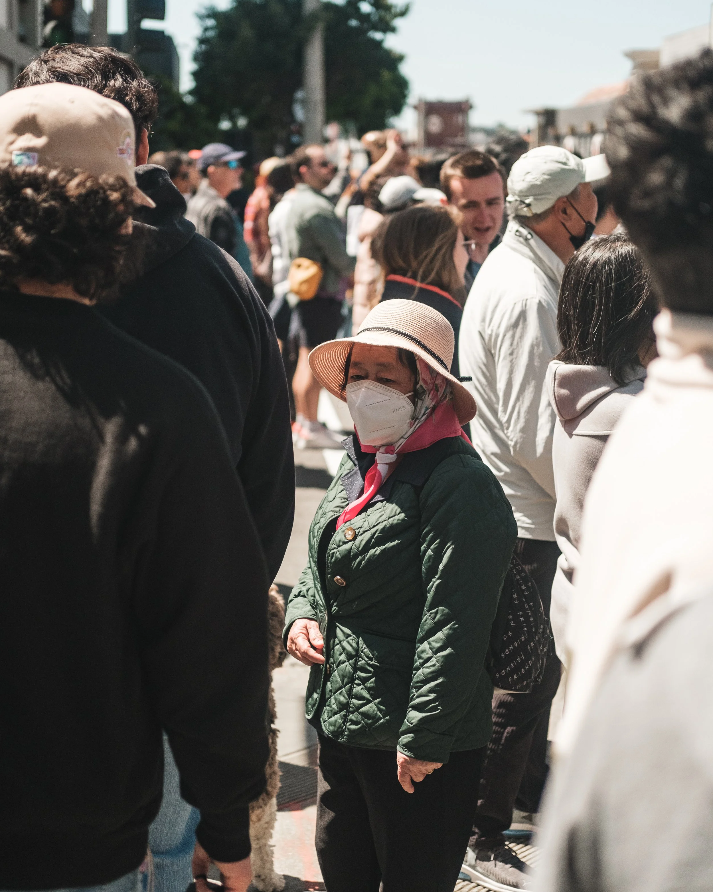 A crowd of people standing outdoors, some wearing masks, with a woman in the center wearing a wide-brimmed hat, face mask, and a green quilted jacket.