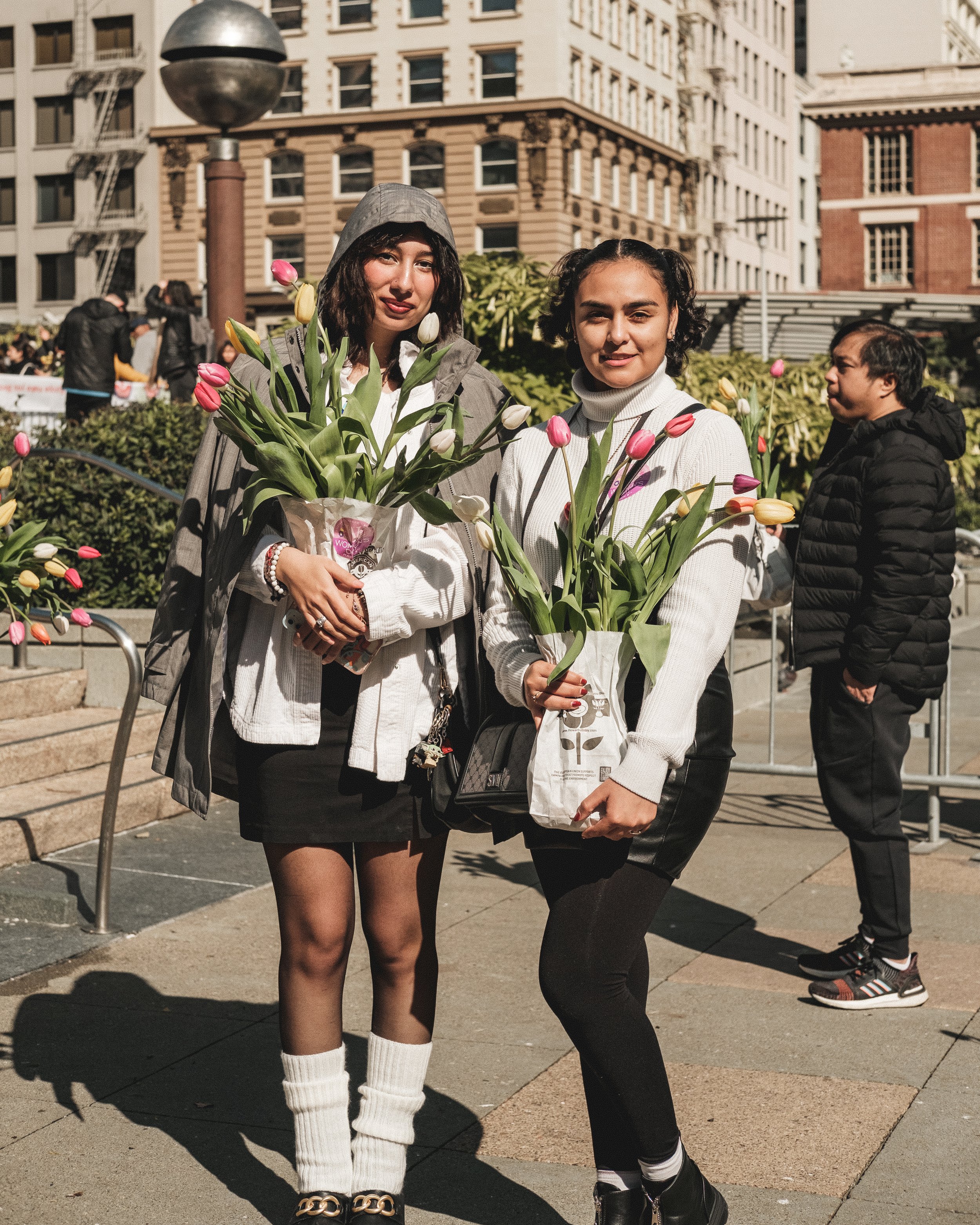 Two young women holding bouquets of tulips pose on a city sidewalk during spring, with tall buildings and other people in the background.
