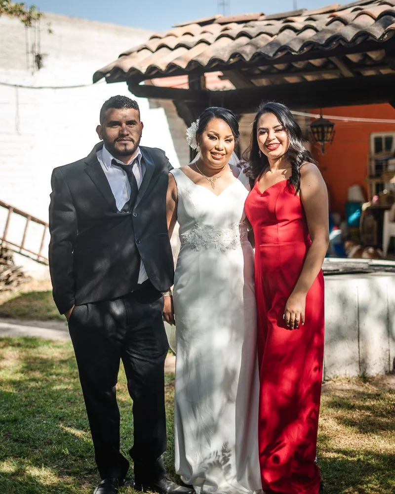 Three people posing outdoors at a wedding or formal event, with a man in a black suit and tie, a woman in a white wedding dress, and a woman in a red dress.