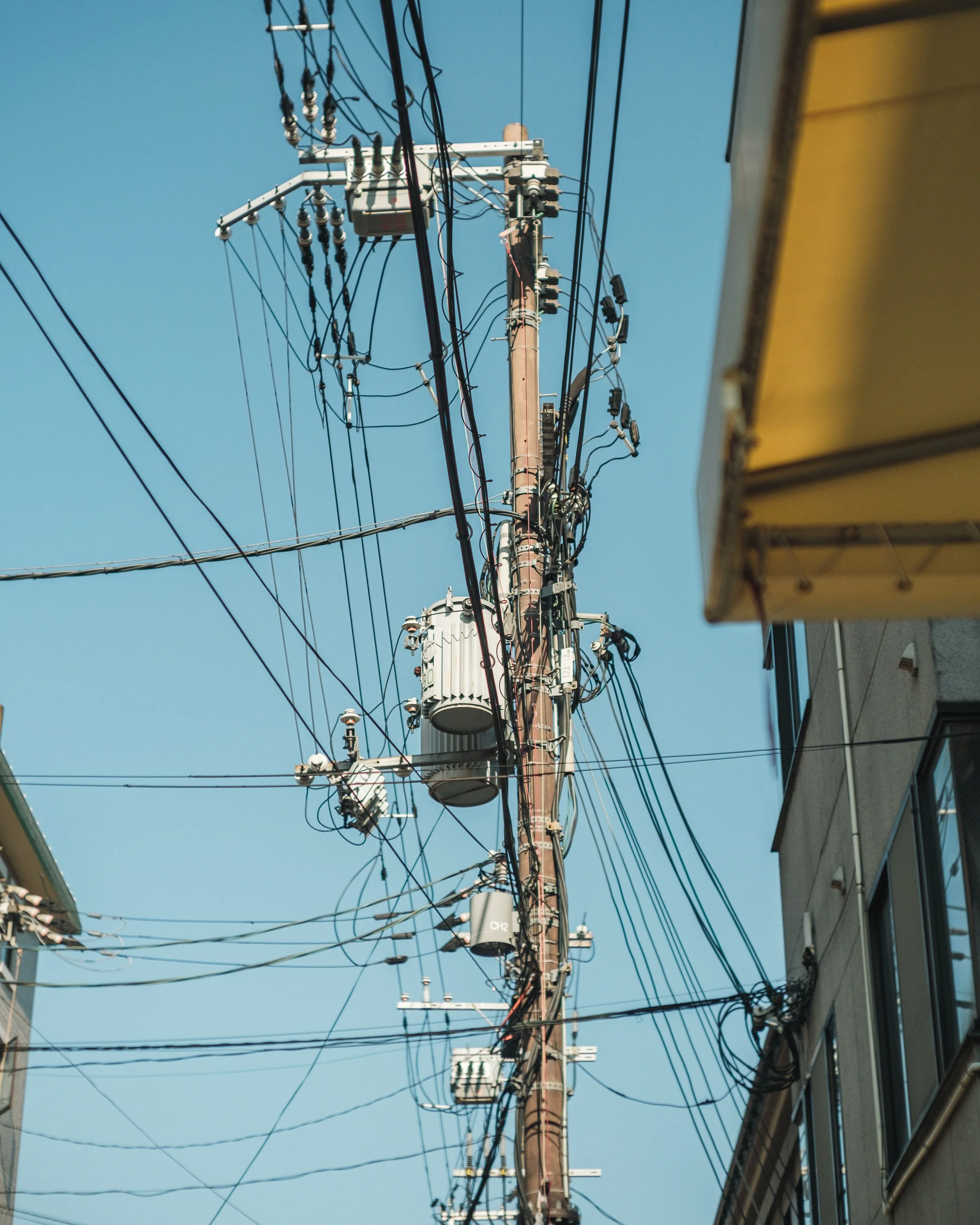 A tall utility pole with multiple electrical wires and transformers, with neighboring buildings and a clear blue sky in the background.