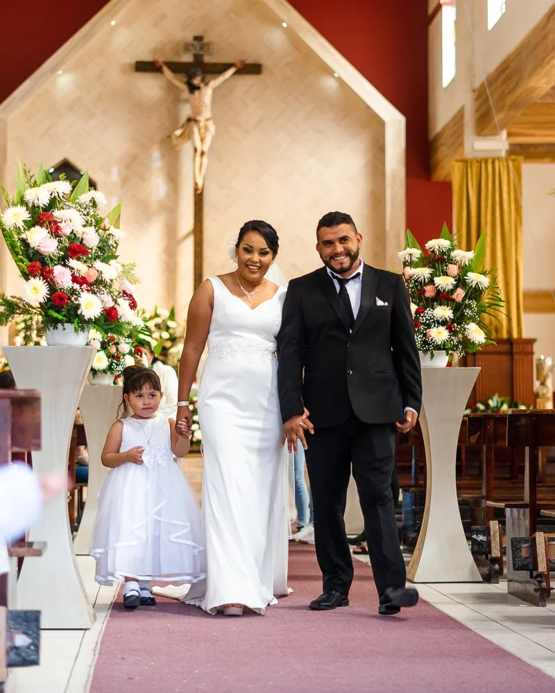 A newlywed couple and a young girl walking down the aisle inside a church, with large floral arrangements on tall stands, a crucifix on the wall behind them, and wooden pews on either side.