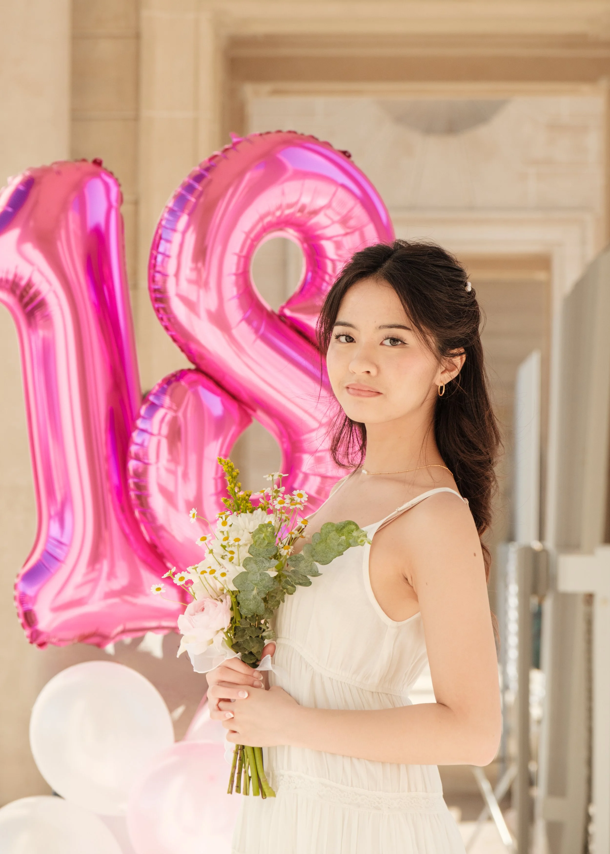 A young woman in a white dress holding a bouquet of flowers, standing in front of pink balloons shaped as the number 18.