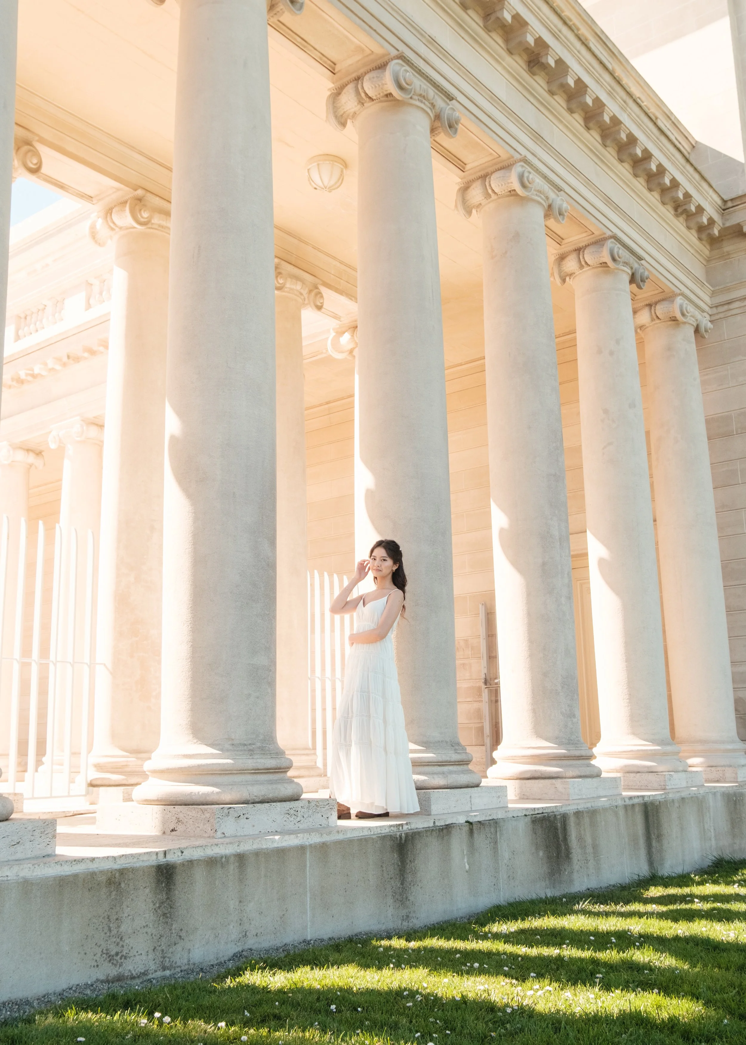 A woman in a white dress standing between tall classical columns outside a building.