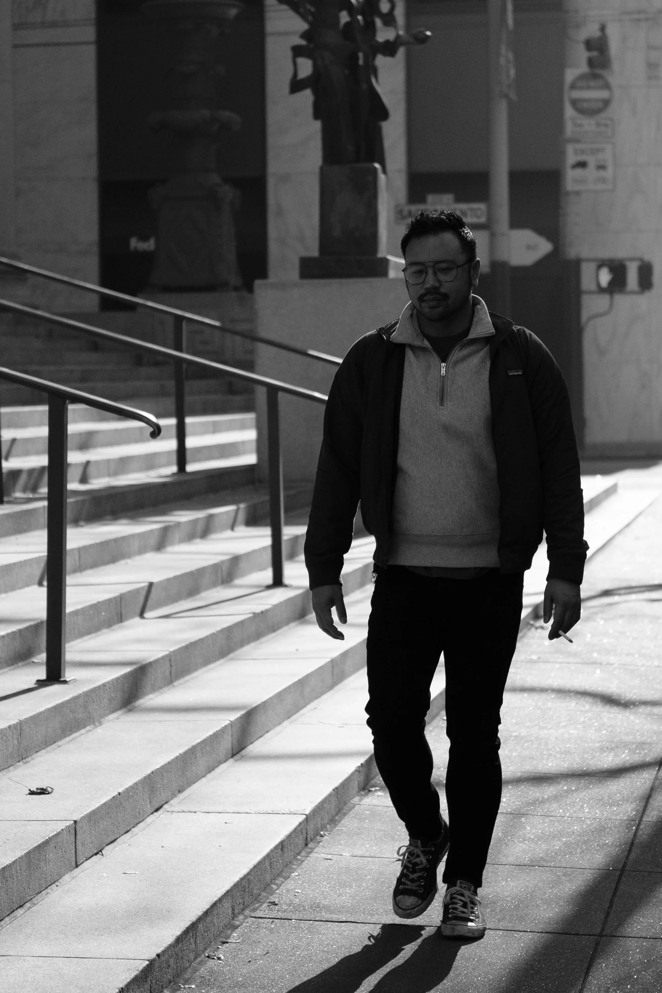 A man walking down a city sidewalk at night, wearing glasses, a jacket, and sneakers, with stairs and street signs in the background.
