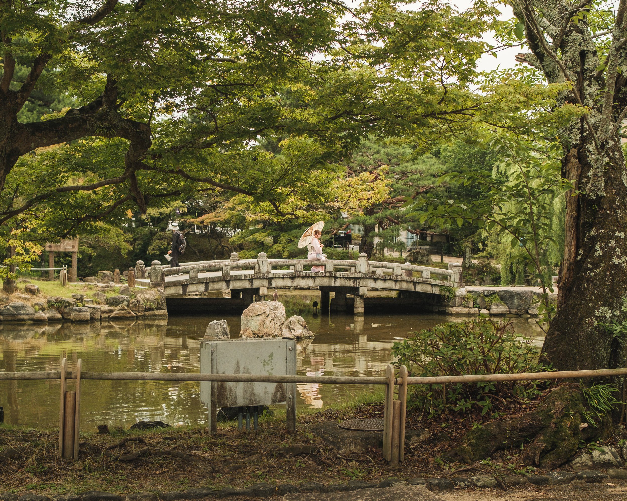 A serene park scene featuring a small stone bridge over a pond, surrounded by lush green trees, with a woman walking across the bridge holding an umbrella, and another person walking nearby.