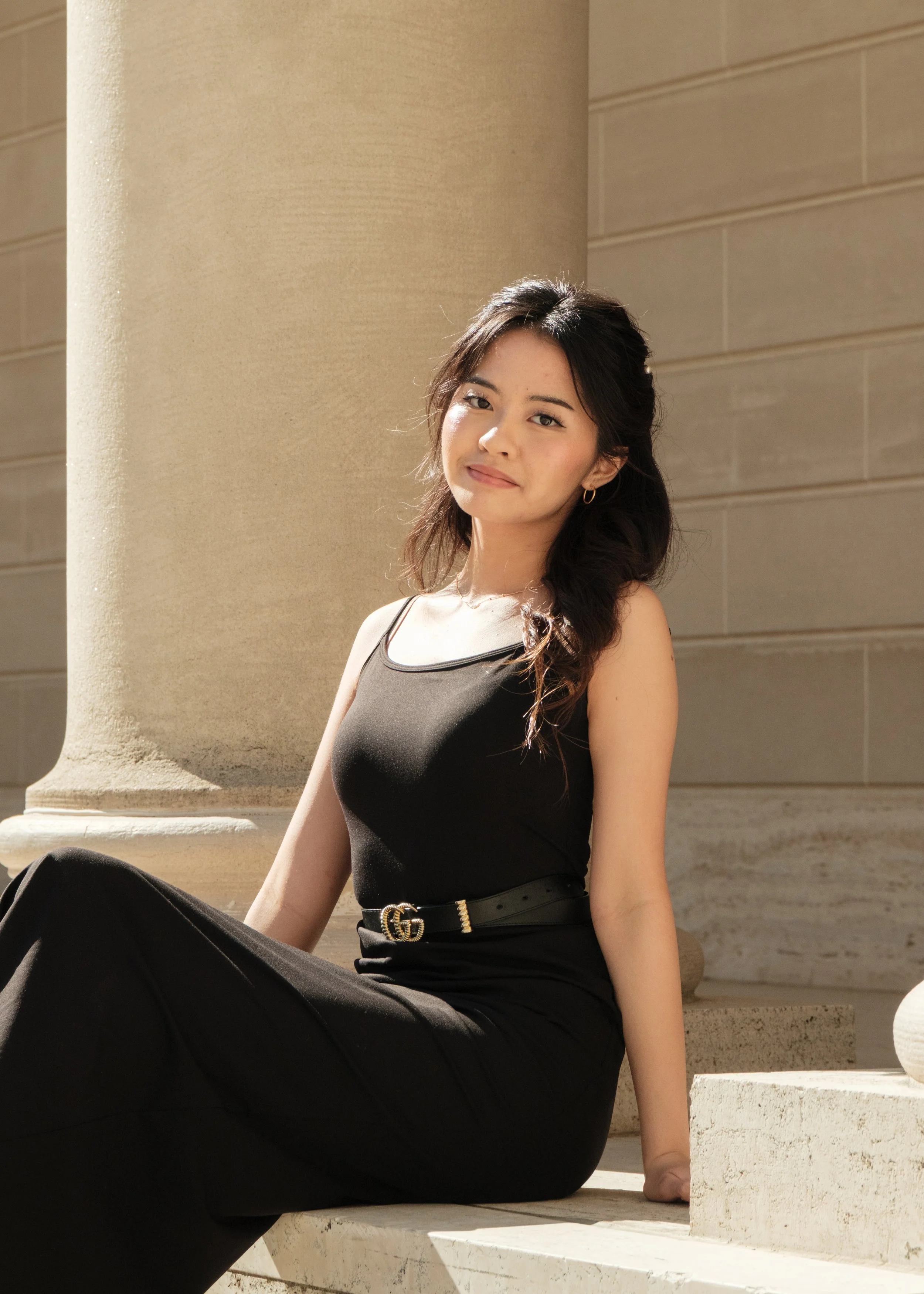 A woman sitting outdoors against a column and stone building, wearing a black sleeveless top and black pants, smiling slightly at the camera.