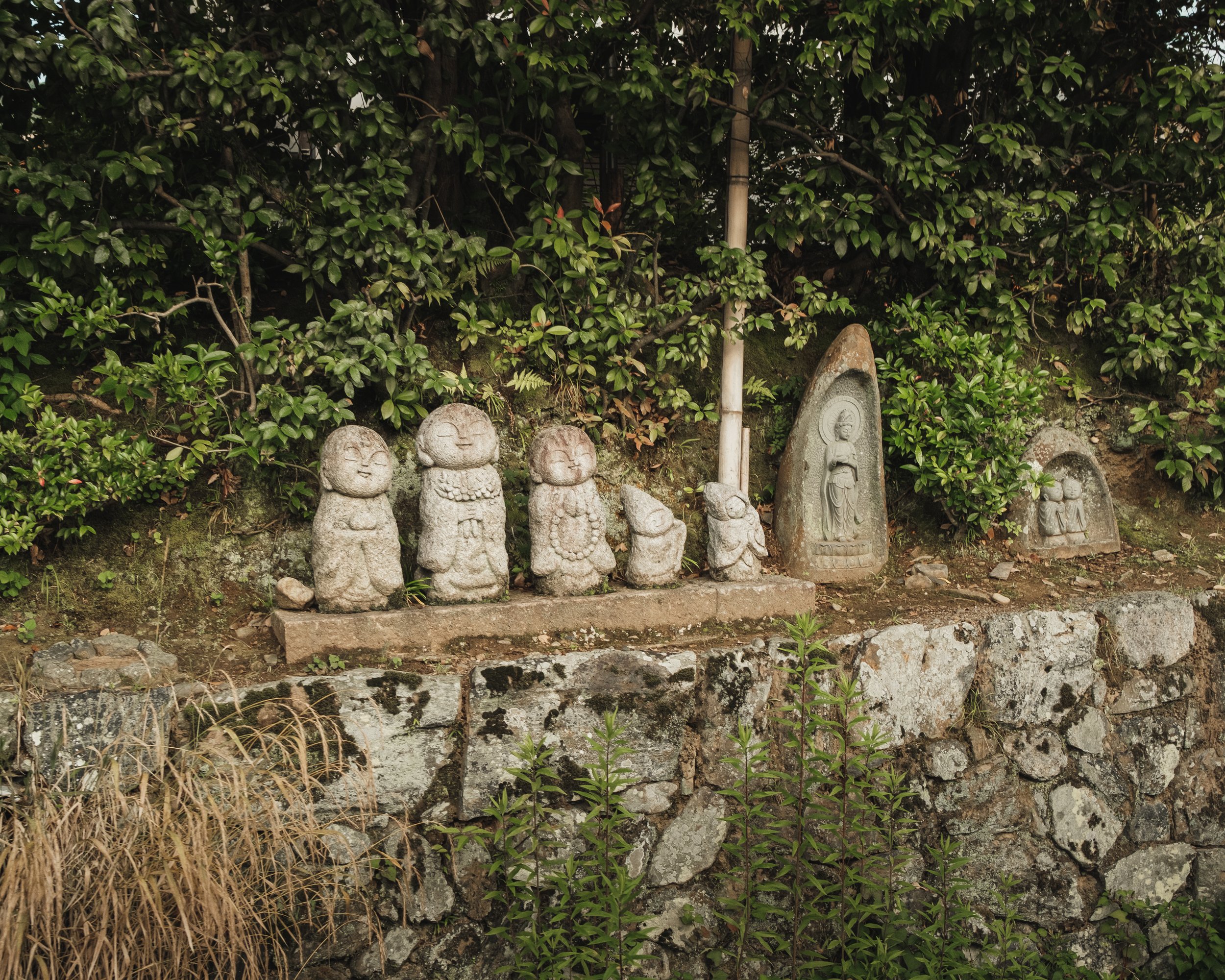 Stone sculptures of smiling Buddhist or spiritual figures and statues, set against a green bushy background on a stone wall.