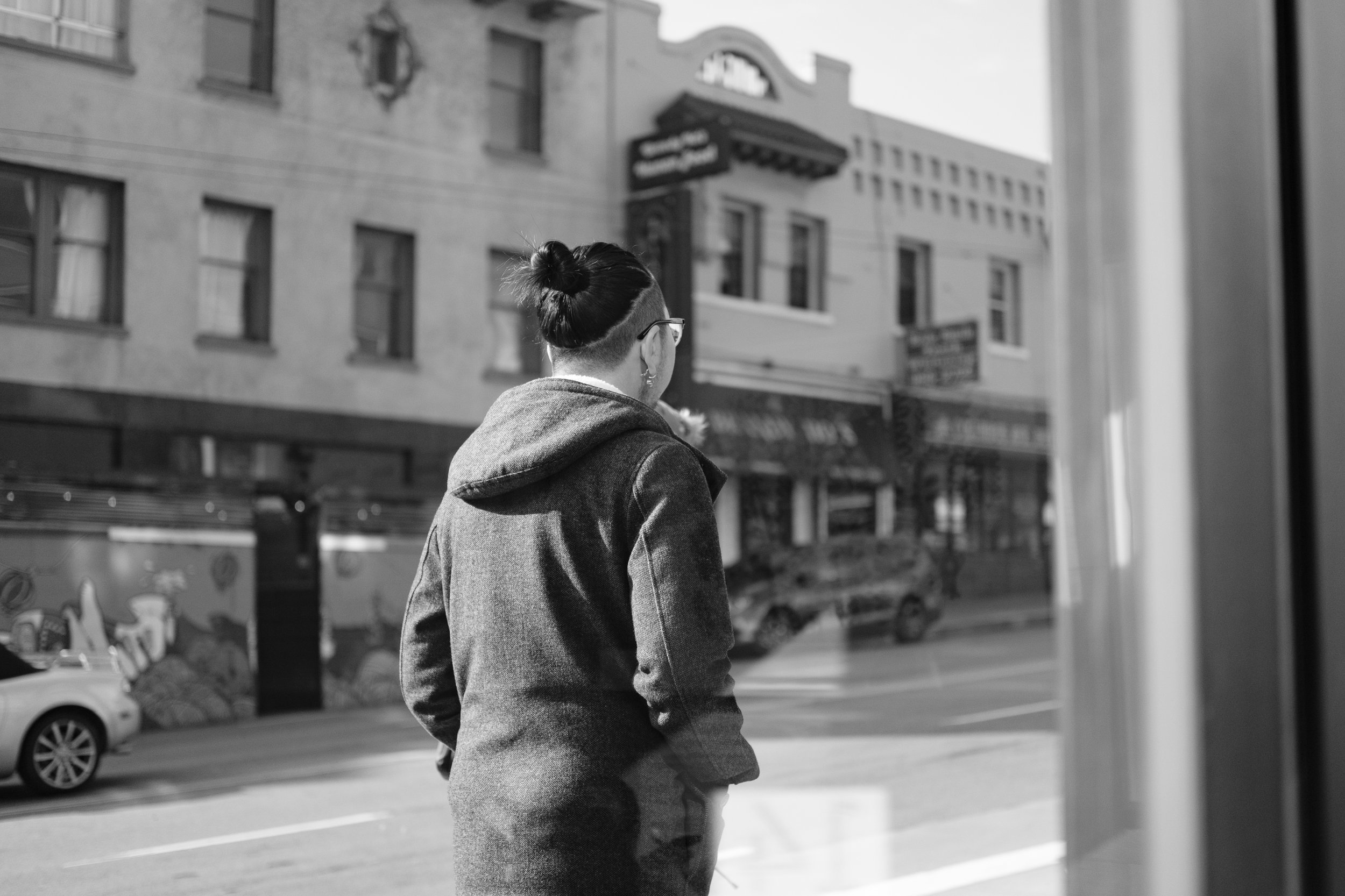 A woman with glasses and a bun hairstyle stands outside, looking at the street through a window, in a city with multi-story buildings, parked cars, and a mural on a wall.