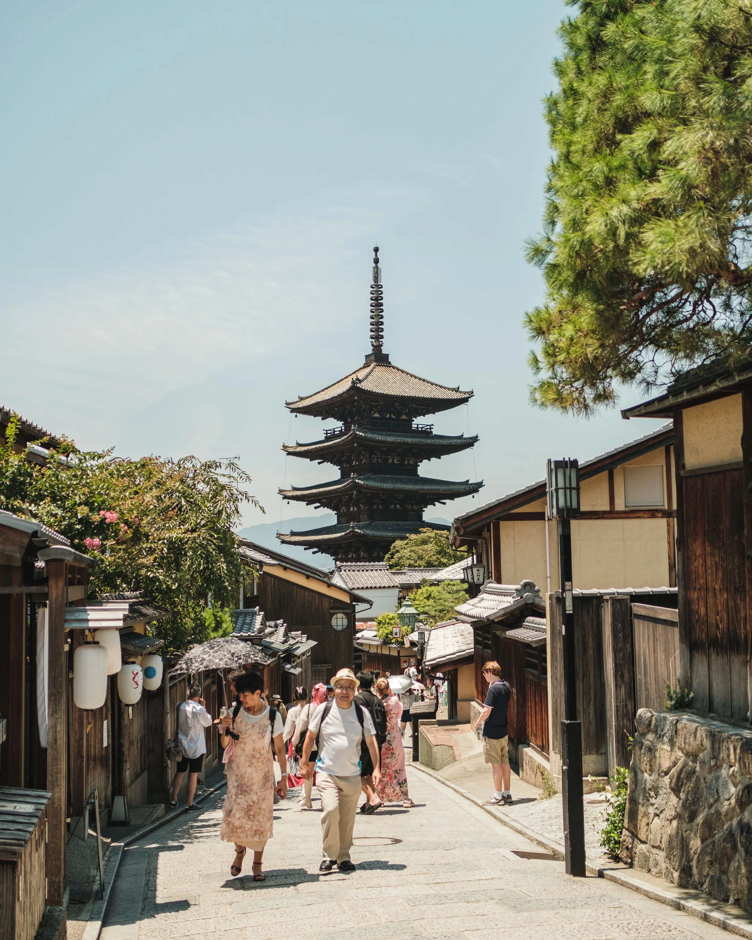 Traditional street scene with wooden buildings, trees, and a five-story pagoda in the background, along with tourists walking and exploring.