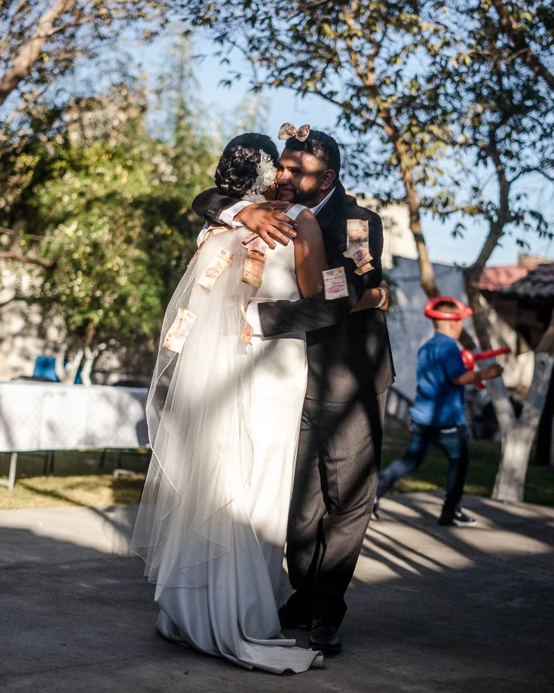 A bride and groom share a hug outdoors during their wedding celebration, with money pinned to their clothing as tradition. A child in the background wears a helmet and plays with a toy.