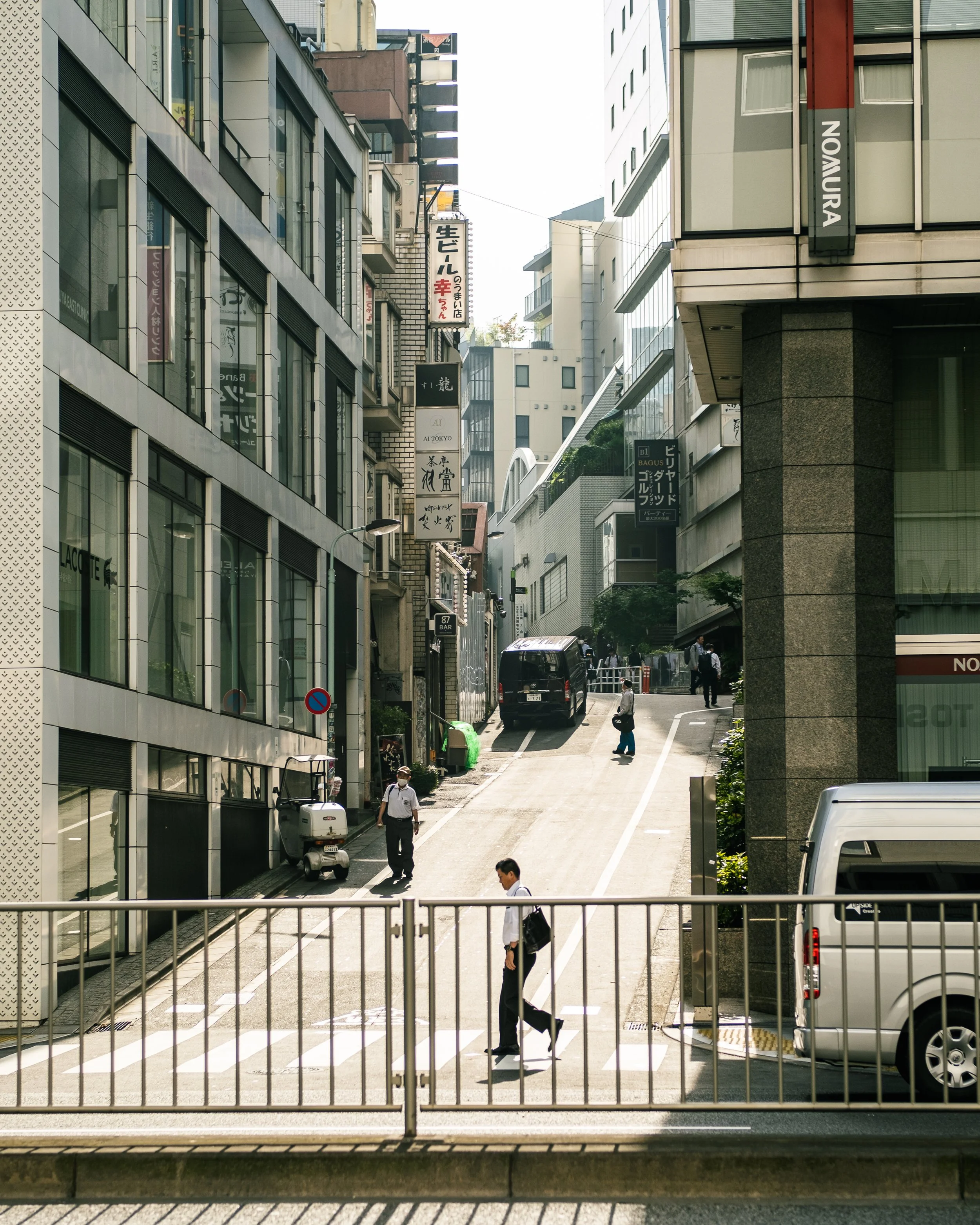 City street scene with buildings on both sides, pedestrians crossing at a crosswalk, and a van parked on the right. Signage in Japanese is visible on the buildings.