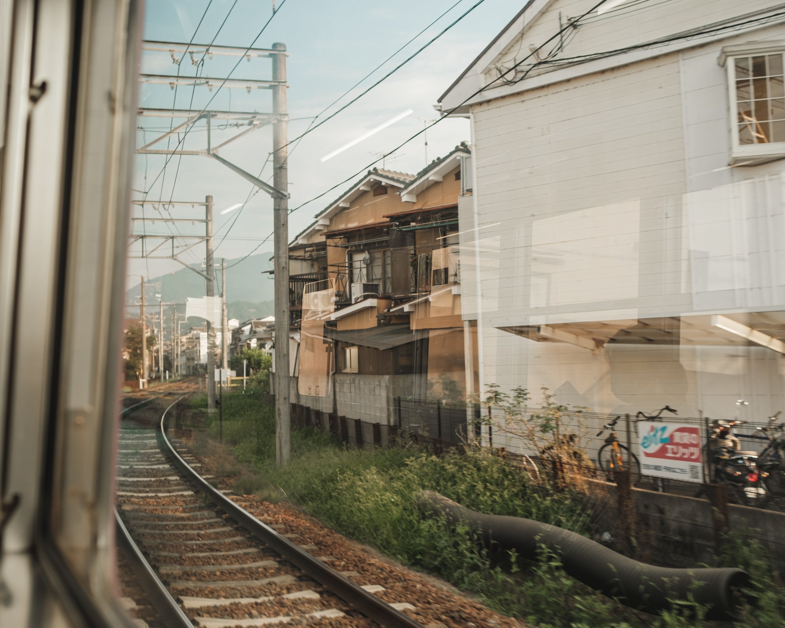 View from a train window showing railway tracks, utility poles, houses, bicycles, and mountains in the background.