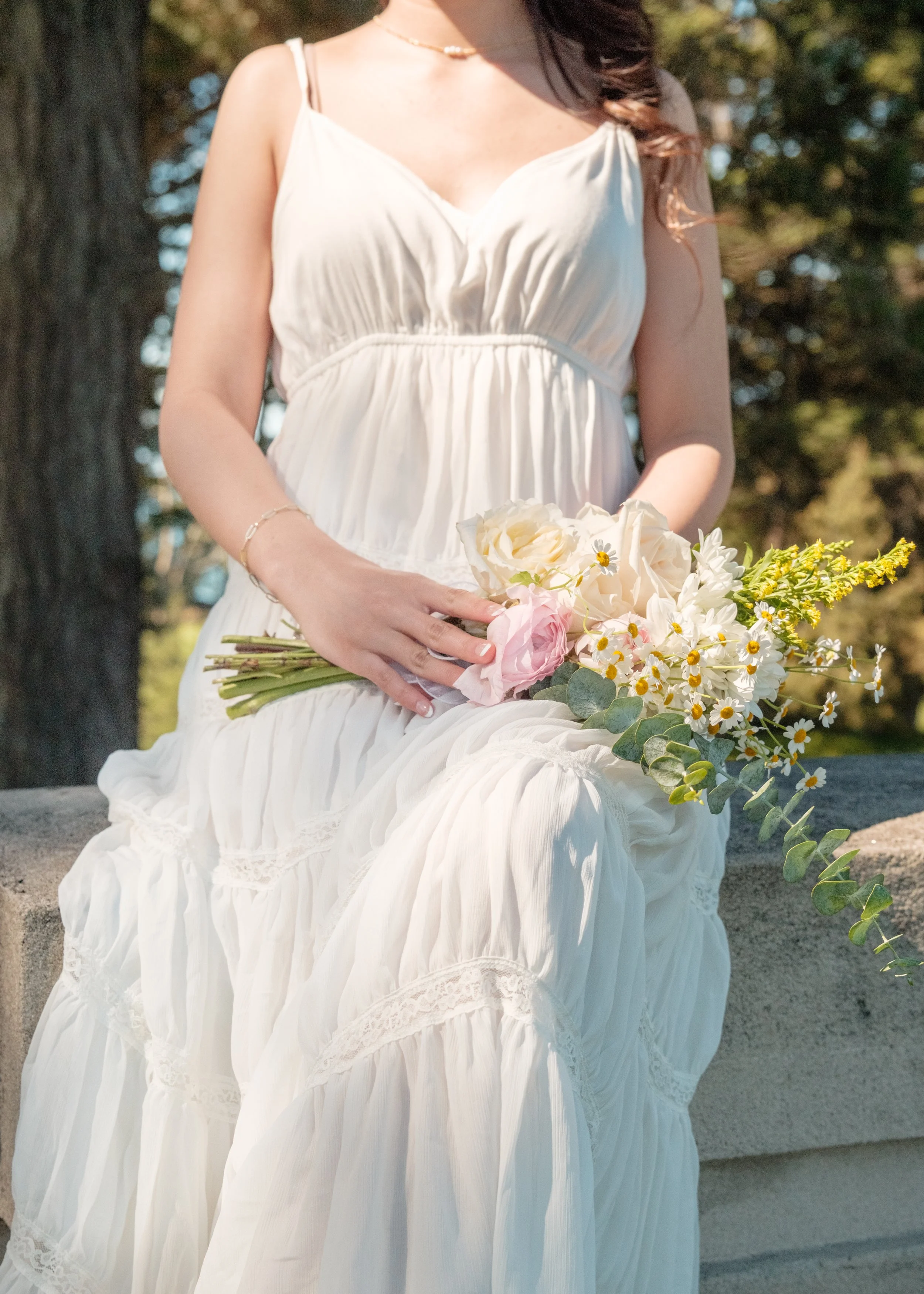 A woman in a cream-colored dress sitting outdoors, holding a bouquet of flowers including roses and daisies.