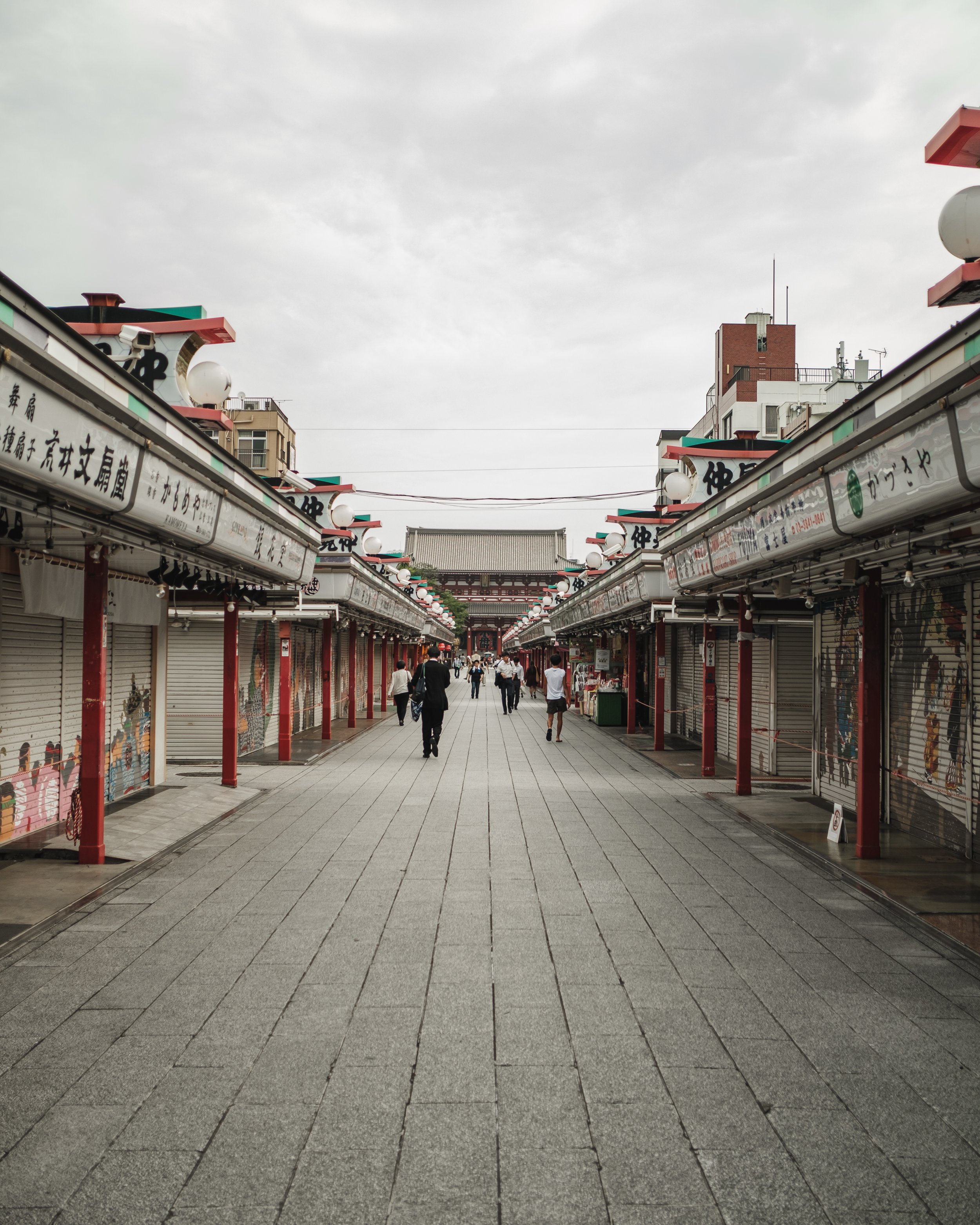 A street lined with closed shop stalls on both sides, with a traditional Japanese building and a torii gate in the background, under a cloudy sky.