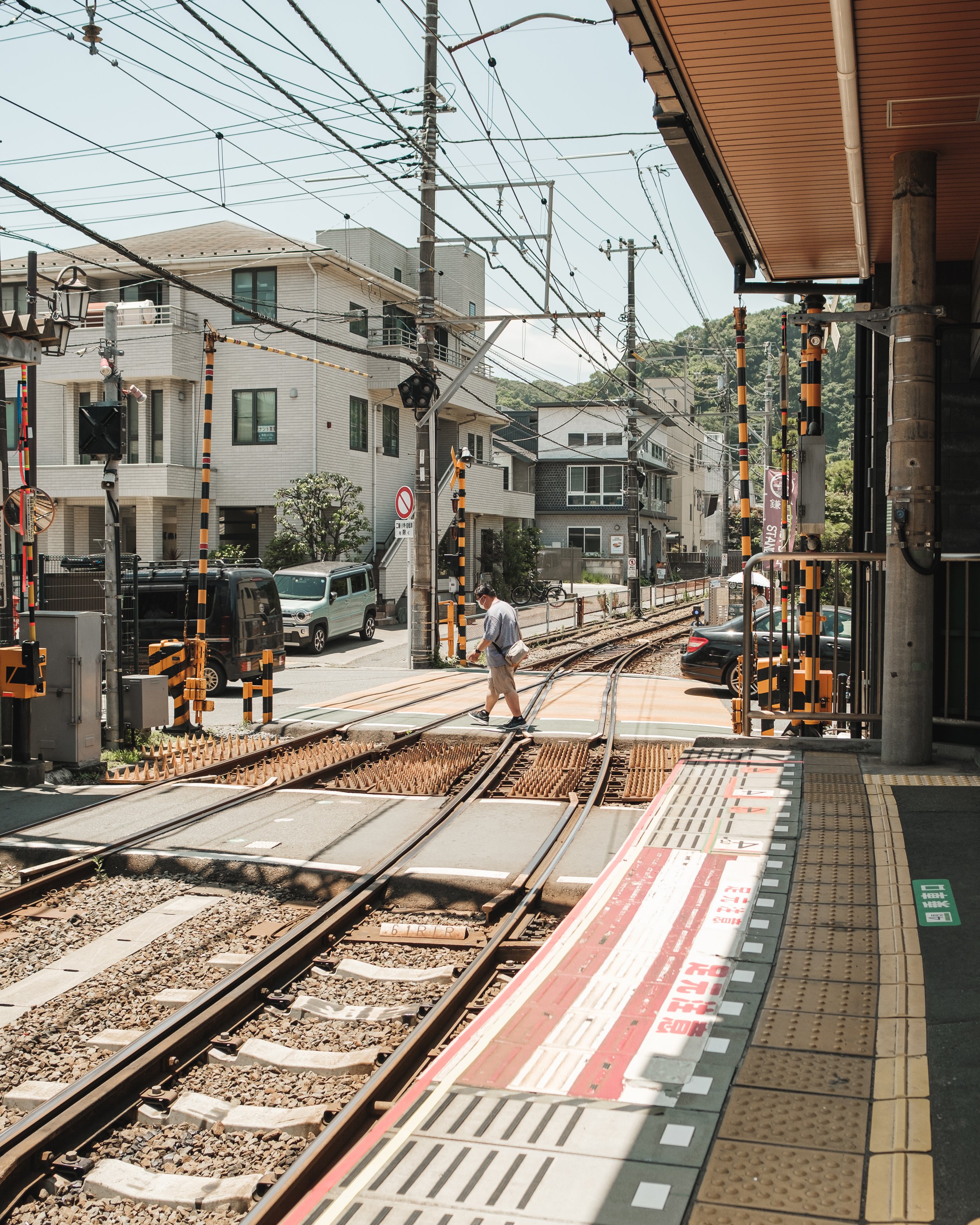 A man crossing railway tracks at a train station platform, with residential buildings and power lines in the background.