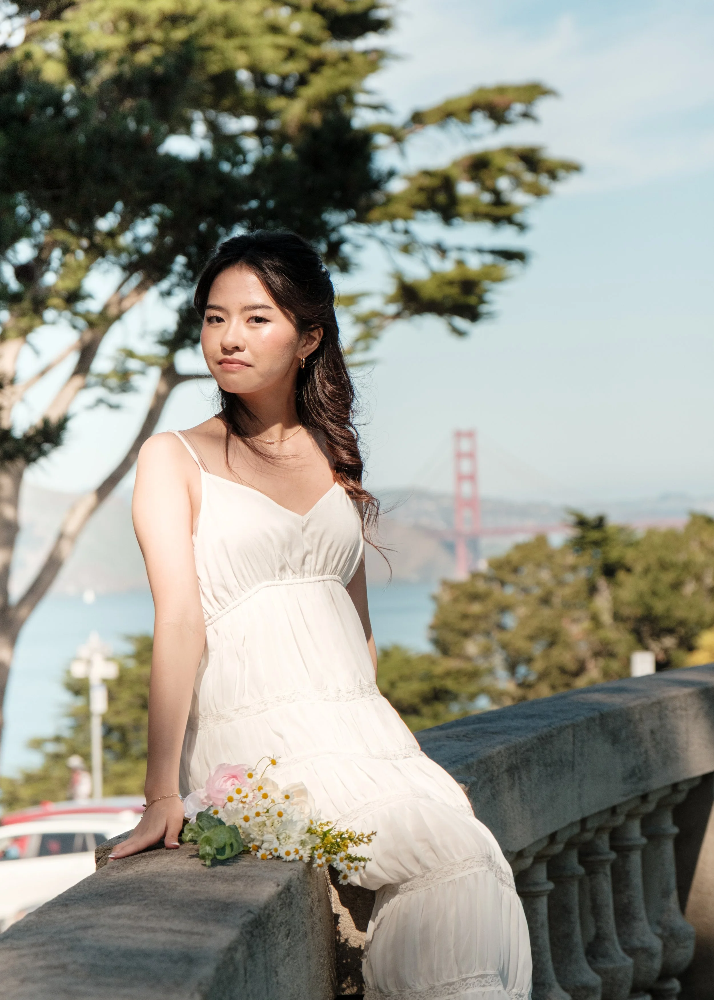 A young woman in a white dress sitting on a stone railing, holding a bouquet of white and pink flowers, with trees and the Golden Gate Bridge in the background.