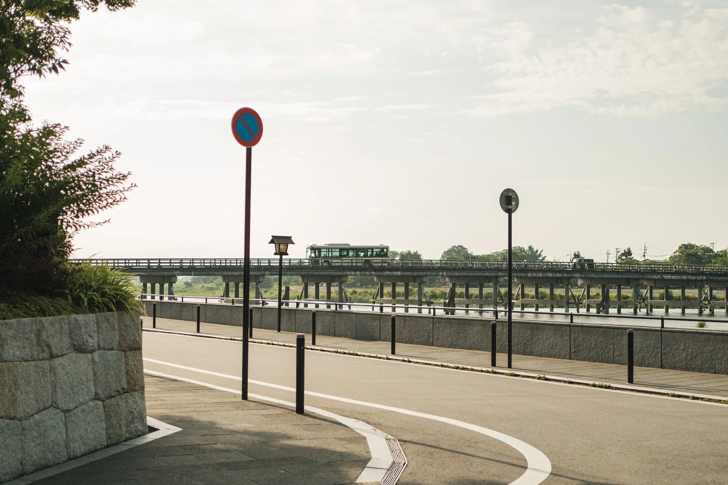 A pedestrian pathway along a waterfront with a no parking sign and a bus on a bridge in the distance.