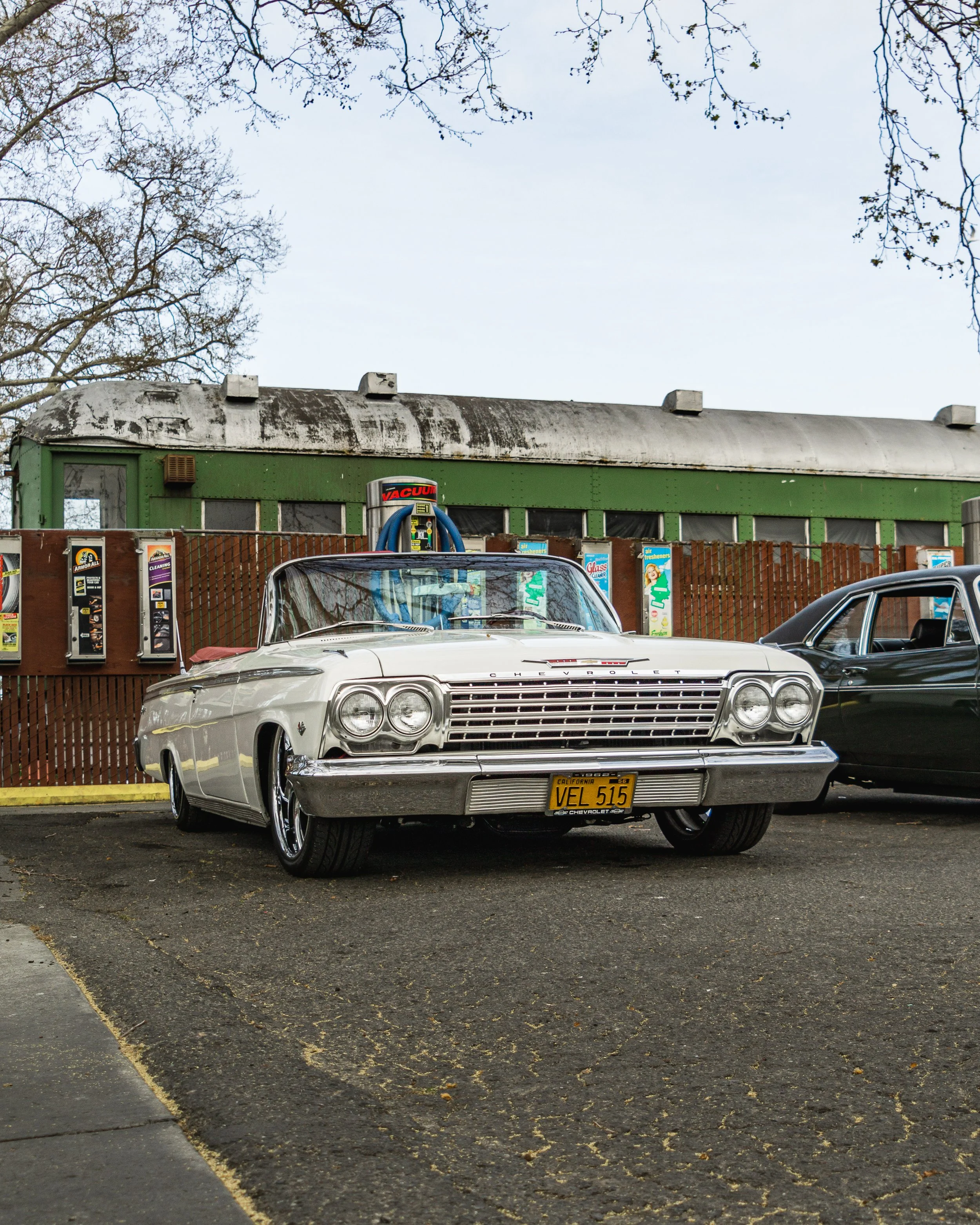 A vintage white Cadillac parked in front of a green building with vending machines, with a black car parked next to it in a parking lot.