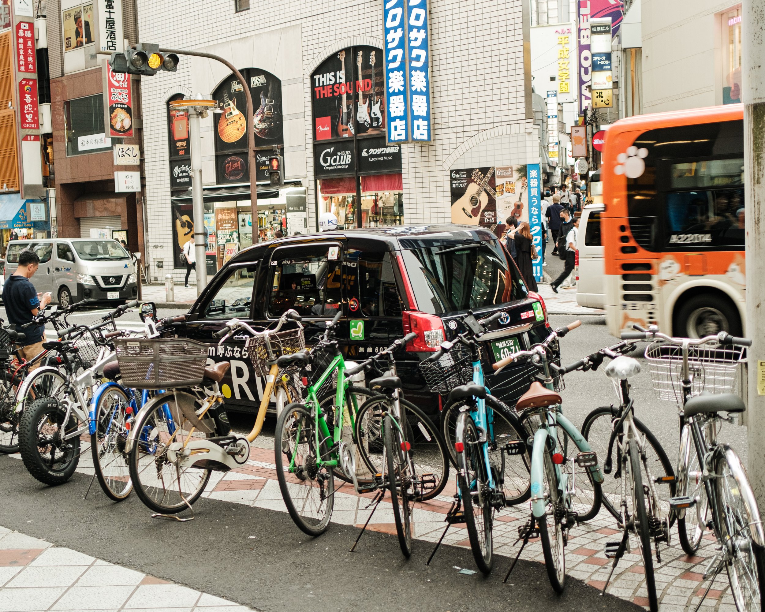 City street scene with bicycles parked along the sidewalk, black taxi, pedestrians walking, and colorful building signs with advertisements and shop fronts.