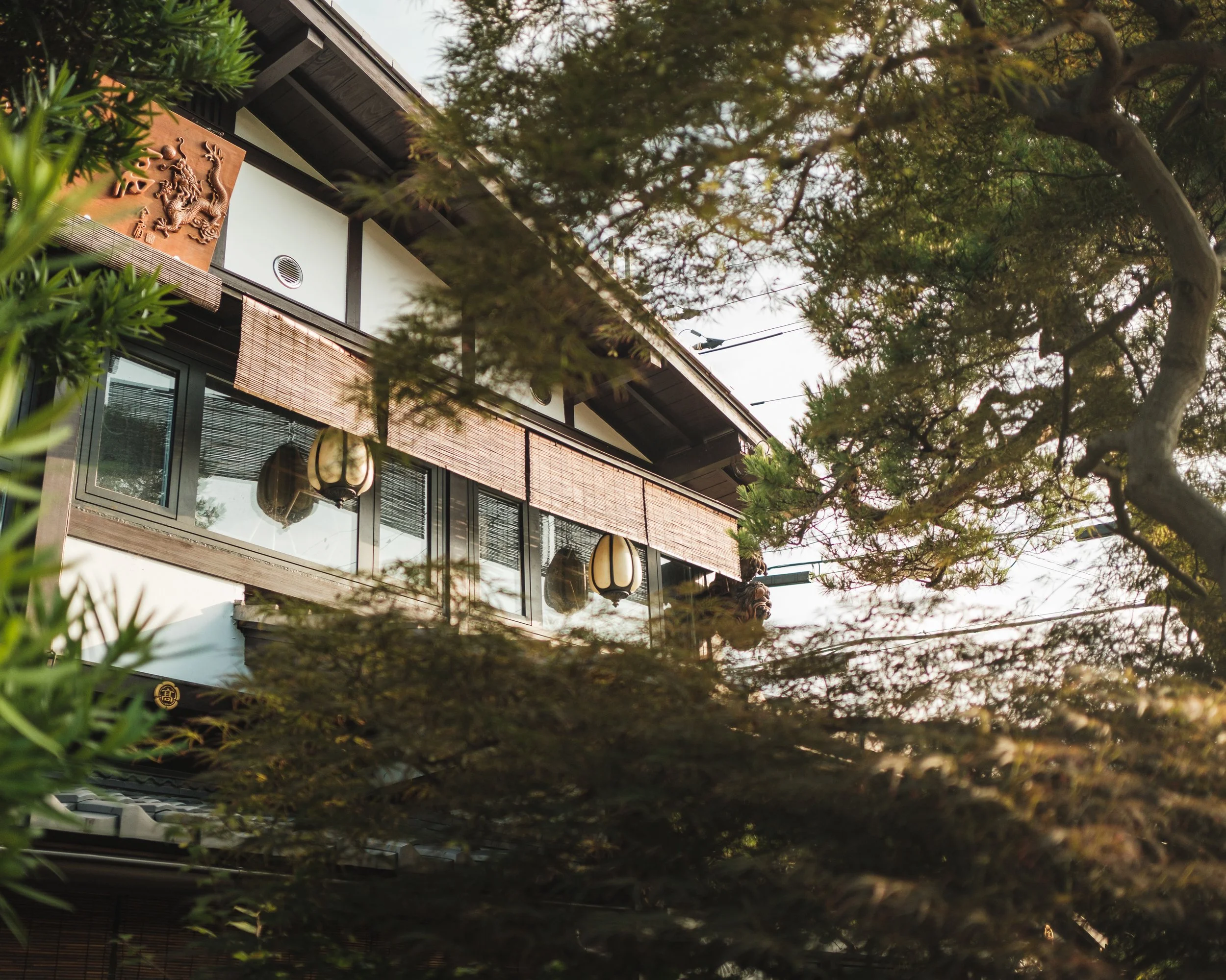 A traditional Japanese building viewed through trees with green foliage, featuring a wooden dragon ornament, paper lanterns, and sliding windows with bamboo shades.