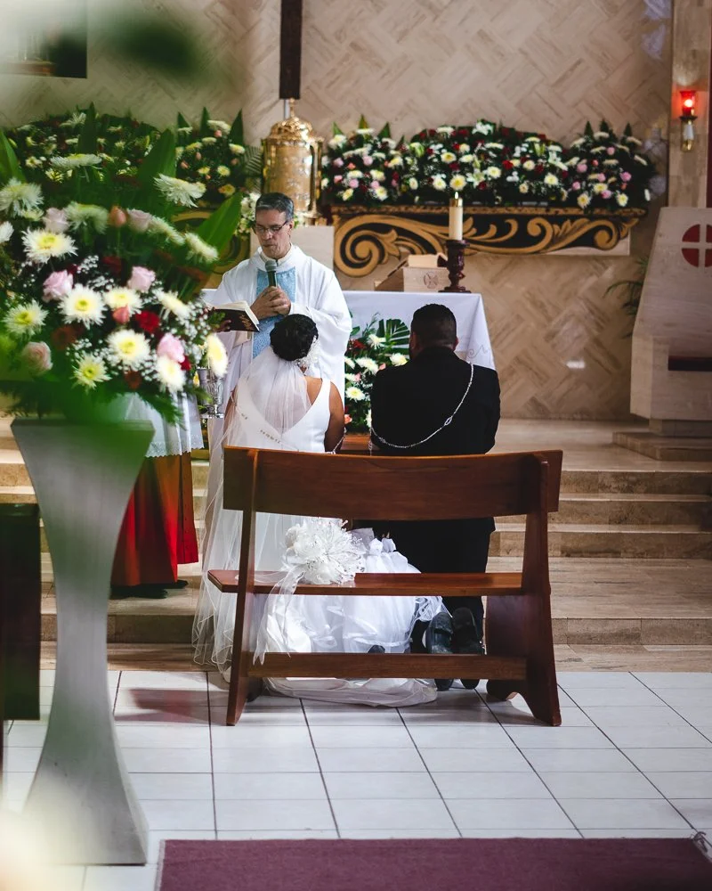 A wedding ceremony with a priest conducting the service, two people kneeling on a wooden bench, and a large floral arrangement in the foreground.